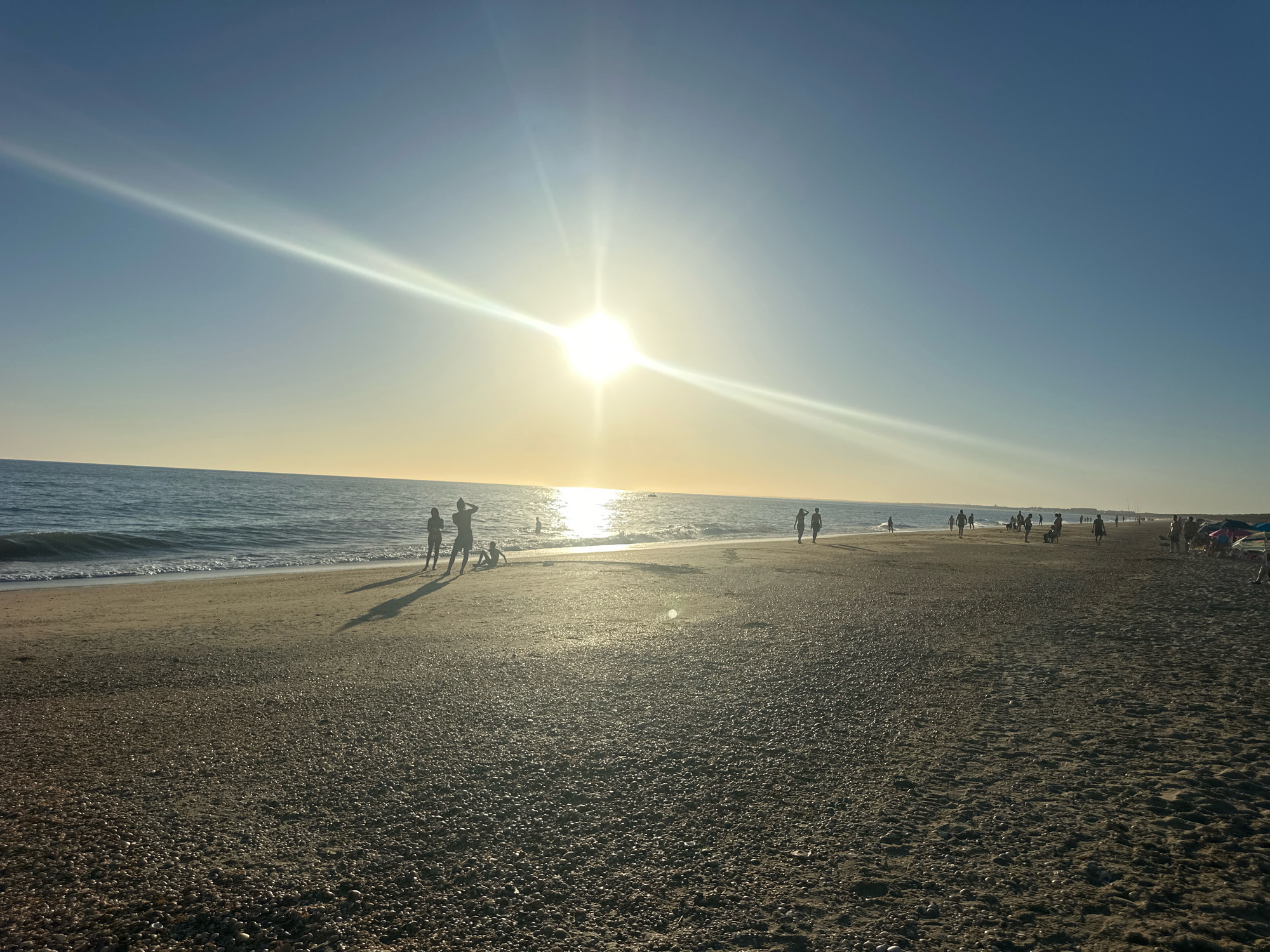 The image depicts a broad, sandy beach at Punta Umbría, Spain, under a clear blue sky with a brilliant sun at its zenith, casting a strong glare and lens flare. The sun's rays reflect intensely on the calm sea. The beach is populated by several individuals, some standing near the water's edge, others walking along the shore, and a few are seated or playing on the sand. The sand itself is a mix of fine particles and small shells, creating a textured surface. The shoreline stretches into the distance, with more people visible further down the beach. The overall atmosphere is one of a typical summer day at the coast.