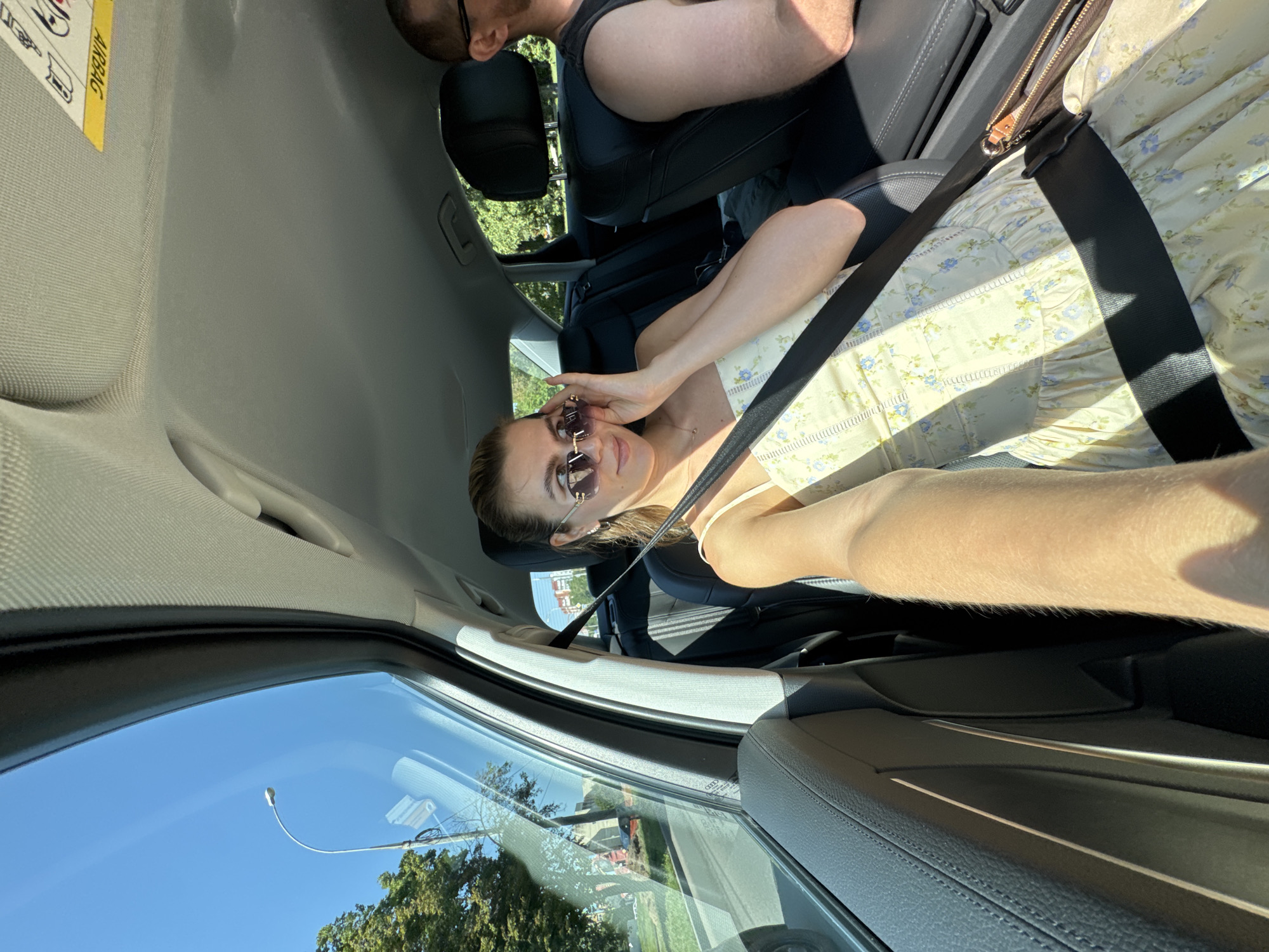 A blonde woman with sunglasses on is in the passenger seat of a car, looking at her hand as she touches her face. She is wearing a light yellow floral dress and a black seatbelt. The car's interior is visible, including black leather seats and the headliner. Outside the car window, a street and trees are visible. A man is in the driver's seat, partially visible on the right side of the image.