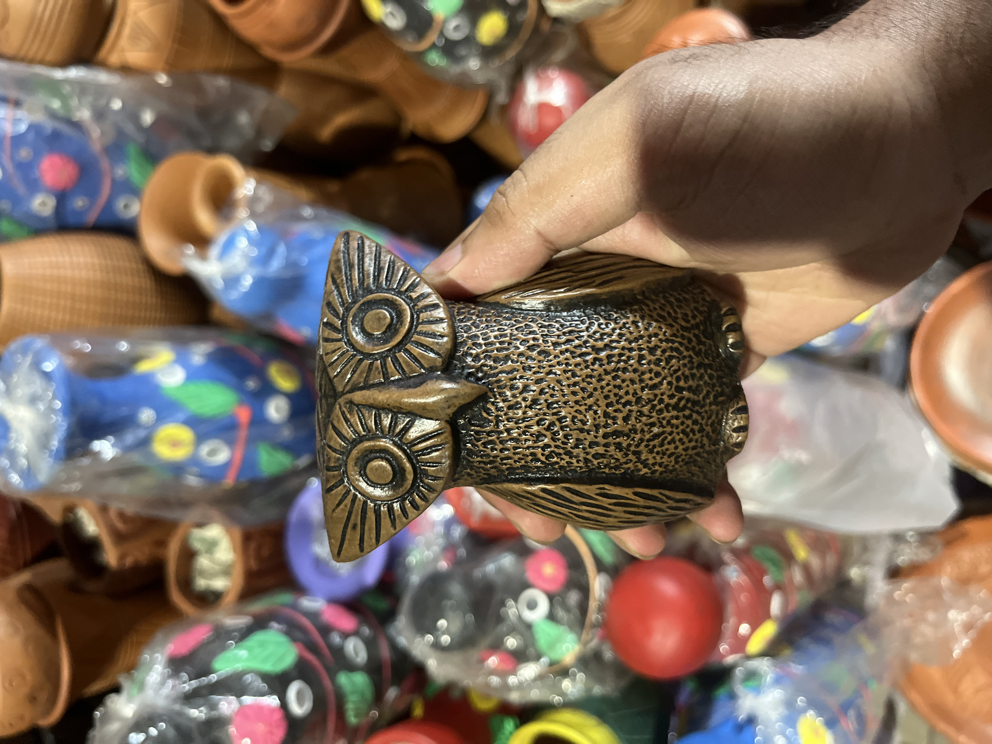 A hand holds a small, brown, owl-shaped handicraft. The owl is made of textured material with stylized eyes. The background is filled with a variety of pottery, some in plastic wrapping, and other handcrafted items in various colors. This image was likely taken in Dhaka, Bangladesh, given the style of the handicraft, location of the store, and the presence of traditional pottery and painted items.
