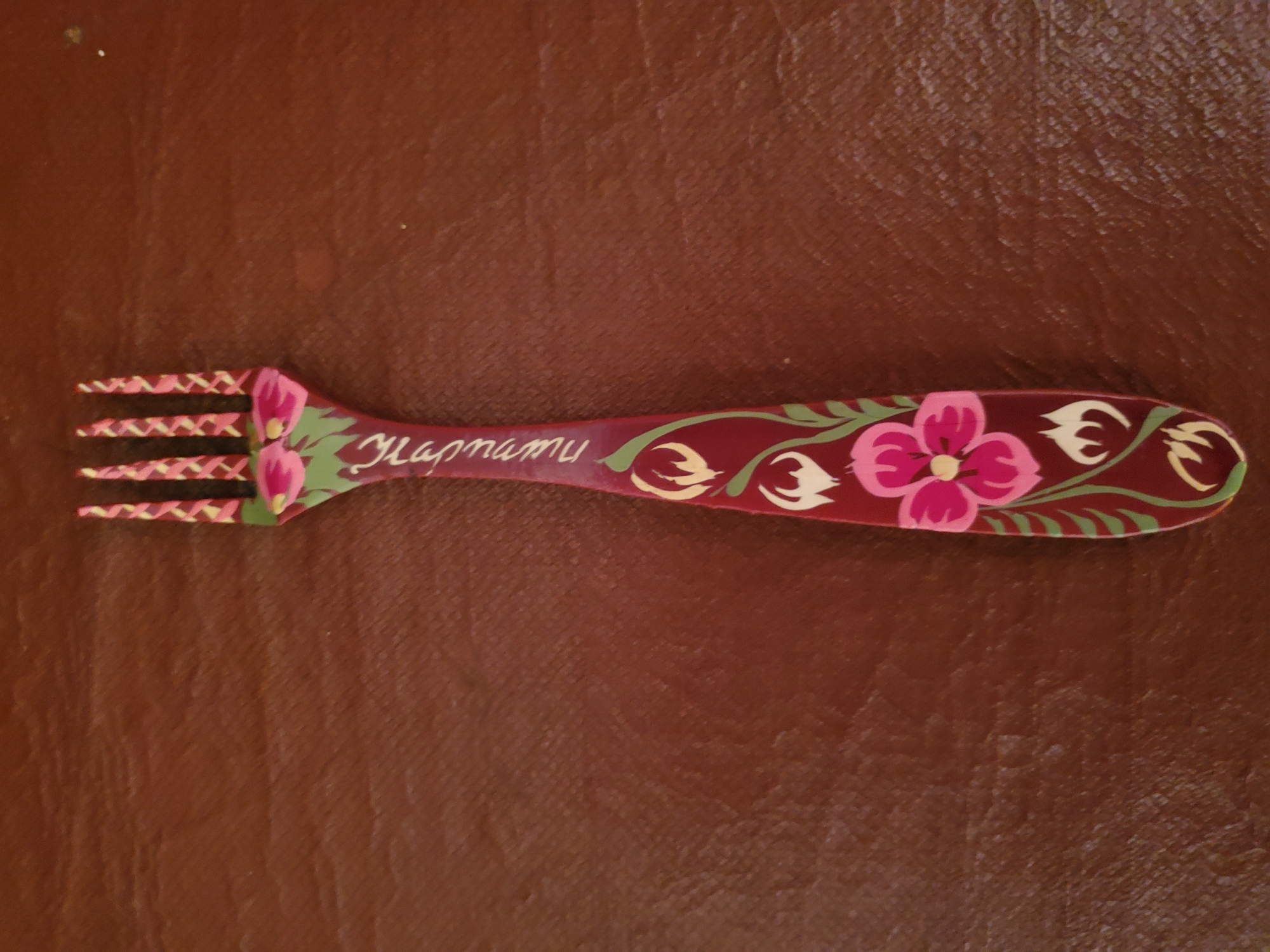 The image shows a close-up, overhead view of a decorative fork lying on a textured, brown surface, possibly faux leather. The fork has a dark red handle painted with a floral design. The tines of the fork are adorned with a pink and yellow crisscross pattern. The handle features pink flowers with green leaves and accents. Inscribed on the handle in white, cursive lettering is the word "Караmu" which appears to be in Ukrainian. The lighting casts subtle shadows on the textured surface. There are no people, actions, or interactions depicted in the image, and no specific location in Konstantinovka, Moldova can be determined from the visual information provided.