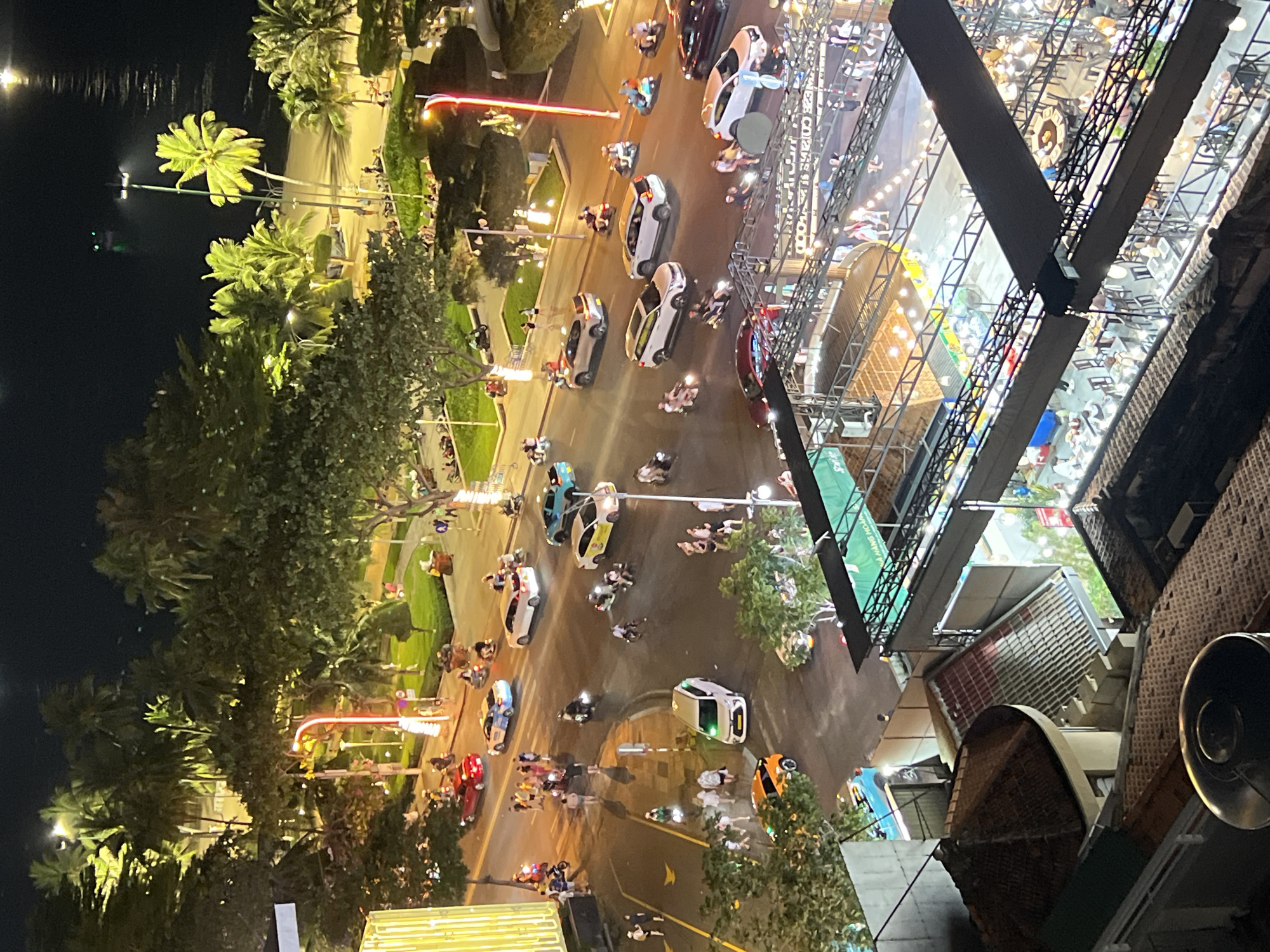 The image captures a nighttime scene from an elevated viewpoint overlooking a street and a beach area in Nha Trang, Vietnam. The foreground features rooftops of buildings, including a circular tiled roof and other structures with visible shingles.
The midground shows a busy street intersection filled with various vehicles. Cars, including sedans and SUVs, are seen driving and parked. Numerous motorbikes with riders are also a prominent feature, indicating common transportation in the area. Pedestrians are walking along the sidewalks and crossing the street. Some individuals are gathered in small groups.
The background reveals a well-lit area with palm trees and green spaces, likely a park or a promenade adjacent to the beach. The beach itself is visible, with the ocean extending into the distance. A small boat with lights can be seen on the water. The overall ambiance suggests a lively urban environment with a mix of residential and commercial activity, along with leisure spaces. The streetlights and building lights illuminate the scene, creating reflections on the wet road surface in some areas.