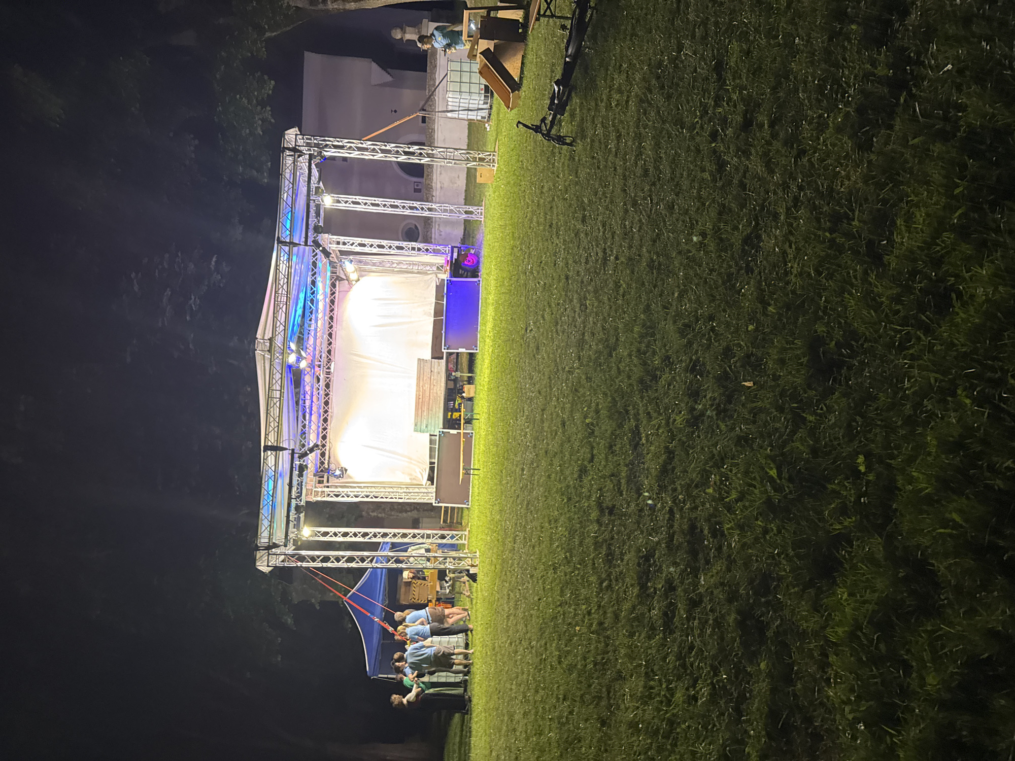 The image captures an outdoor event at dusk or night, likely in Höhenkirchen-Siegertsbrunn, Germany. A stage setup with lighting rigs is the central focus, bathed in a mix of white and blue light. A large white screen forms the backdrop for the stage, suggesting a performance or presentation is about to begin or has just concluded. In the foreground, a group of people, some wearing blue shirts, are gathered near the stage, possibly as performers or crew members. Their casual attire and relaxed postures suggest an informal event. The scene is set on a grassy field, which slopes gently downwards, indicating an outdoor venue like a park or festival grounds. In the background, a building or structure is partially visible, adding to the sense of place. A bicycle is parked near the stage structure, a subtle detail that hints at the mode of transportation for some attendees or organizers. The time of day is clearly night or late evening, as indicated by the darkness and the prominent stage lighting. The grass appears somewhat damp, possibly from recent rain or dew, but the overall atmosphere seems calm and ready for the event. No visible text is present in the image.