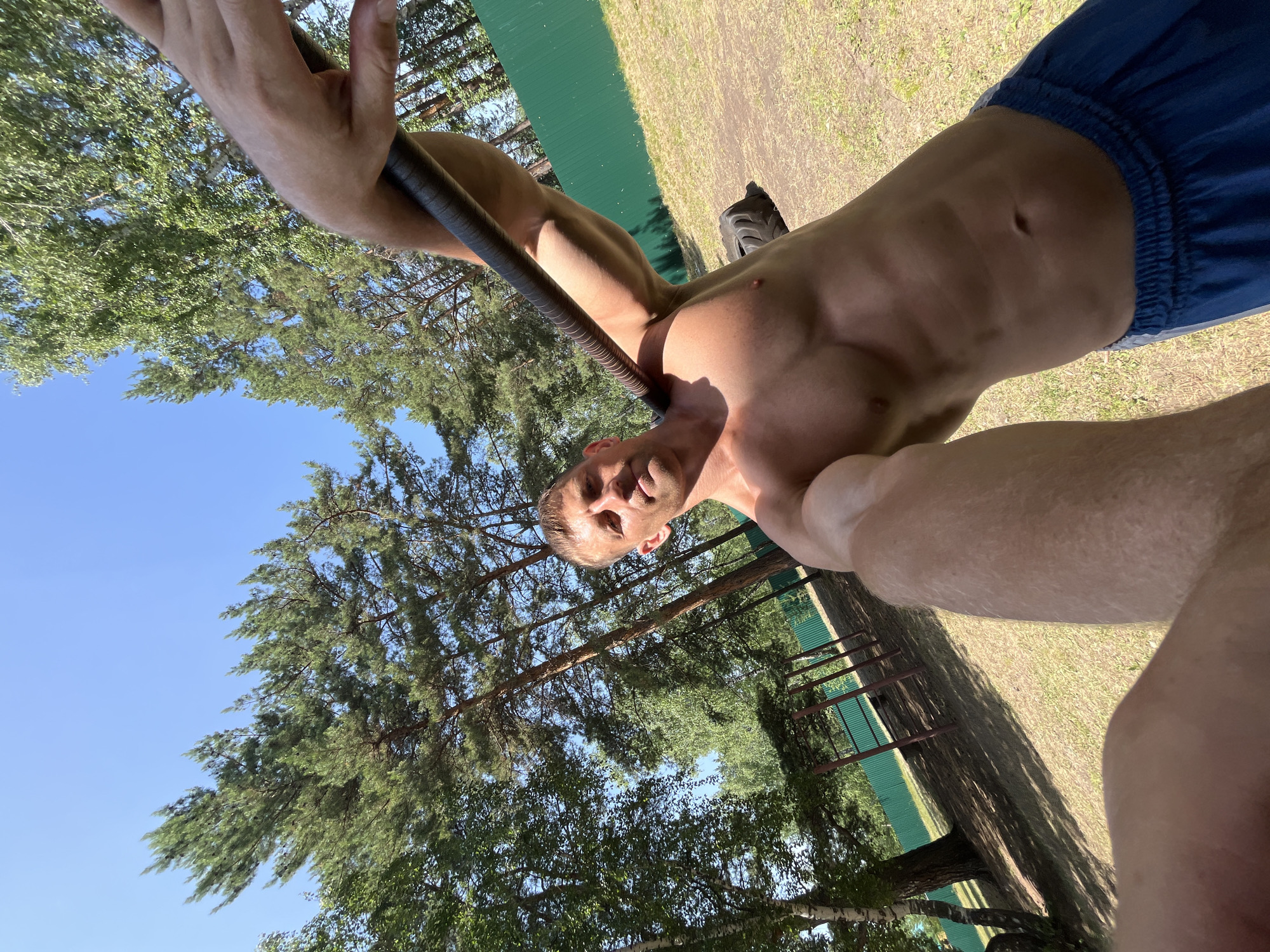 A shirtless man with muscular build is holding a dark-colored bar over his shoulders while looking at the camera. He is outdoors in a grassy area with trees in the background. A green fence is visible behind him. There is also a large black tire on the ground to his right. The sky is clear and blue. The location appears to be an outdoor gym or park, possibly in Krasnoturansk, Russia, given the presence of pine trees and birch trees, which are common in that region.