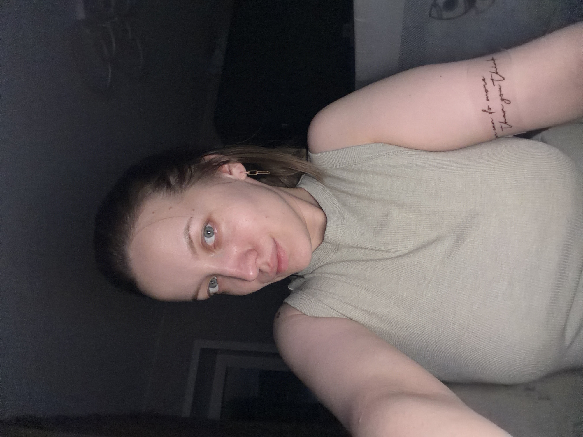 The image captures a young woman taking a selfie, likely indoors in Reshetnikovo, Russia, under dim lighting conditions, suggesting evening or night. The woman has fair skin, light-colored eyes (appearing blue or grey), and light brown hair pulled back from her face. She exhibits a calm and subtle, faint smile.
She is wearing a light, possibly sage green or beige, ribbed short-sleeved top. On her left ear, a small, golden, rectangular hoop earring is visible. A prominent detail is a script tattoo on her right forearm, which reads: "you can do more than you think". The tattoo text wraps around her arm.
The background is dark and largely indistinct, indicating an indoor setting with limited illumination. The focus remains on the woman's face, upper body, and the visible details like her earring and tattoo. Her expression is serene.
