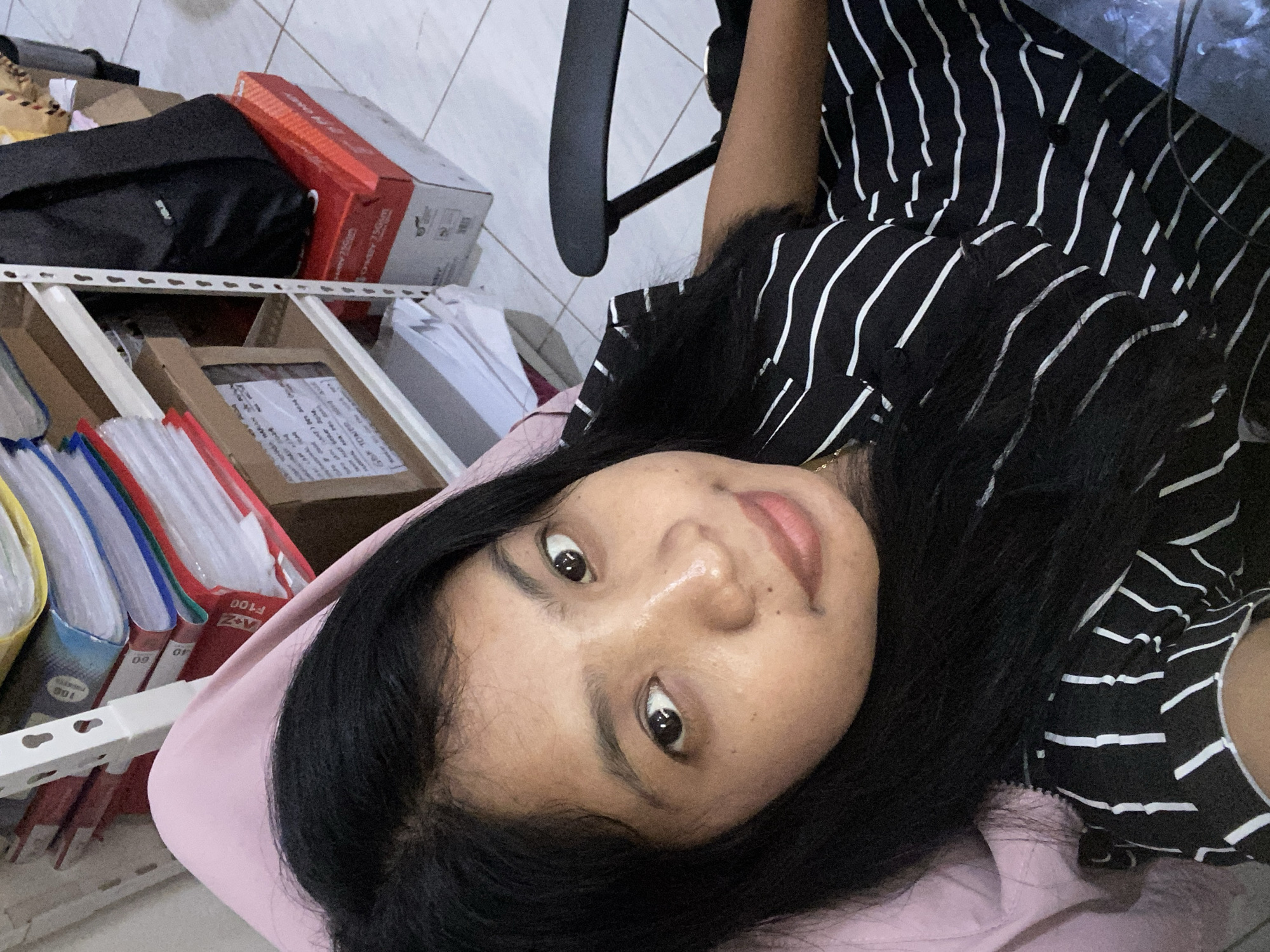 The image is a close-up selfie of a person with dark hair and tan skin, wearing a black and white striped shirt. The person is looking at the camera with a slight smile. The background is a cluttered workspace with shelves containing files, boxes, and other items. The environment appears to be indoors with tiled walls. The location is potentially Negara, Indonesia.