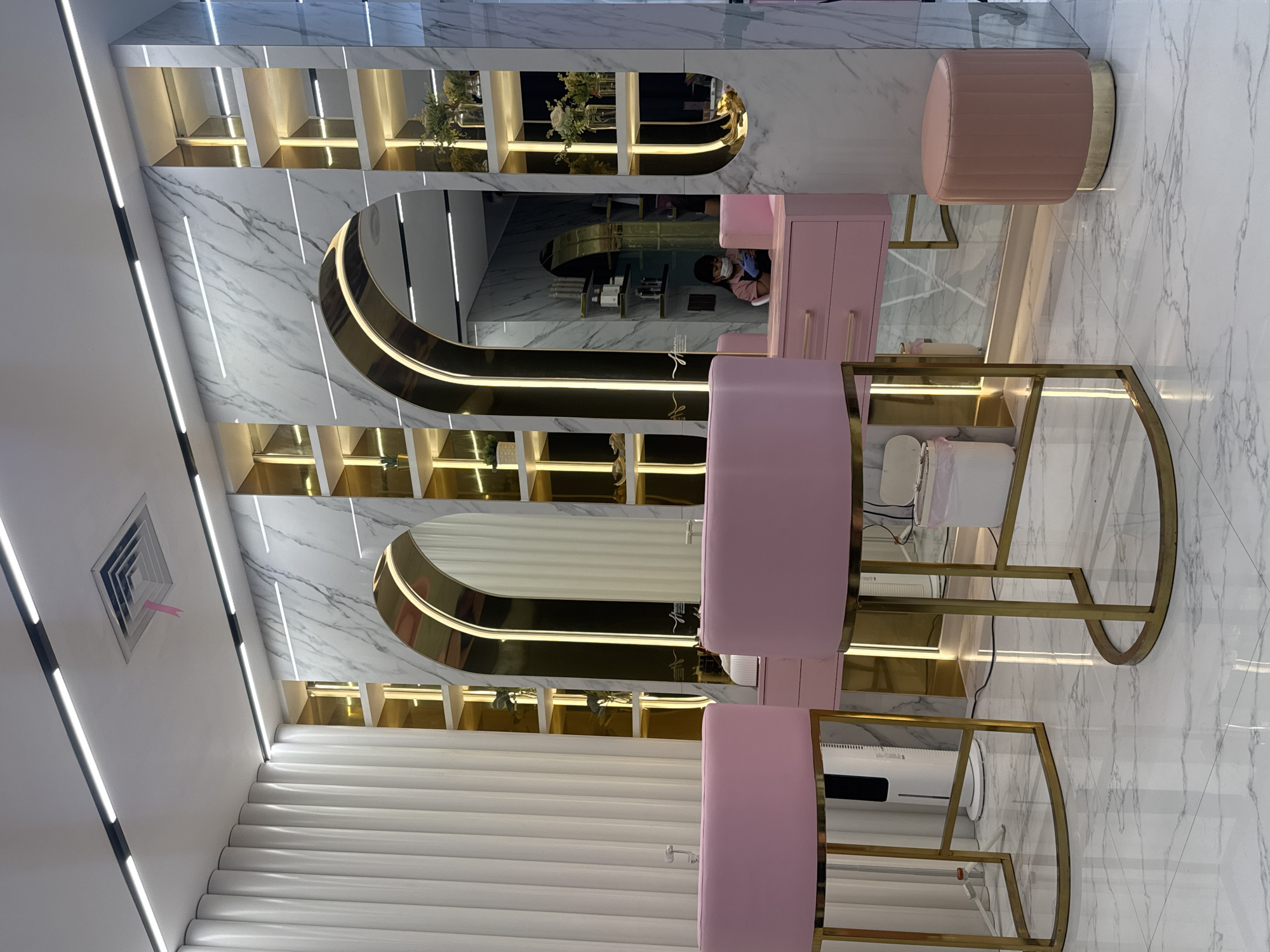 This is an interior shot of a commercial space in North Kuta, Indonesia, possibly a nail salon or beauty parlor. The decor is modern and luxurious, with a pink, gold, and marble color scheme. There are multiple manicure stations with pink upholstered seating and gold metal frames. Mirrored areas with gold arched borders and built-in shelving are visible, some containing decorative plants. A person wearing a face mask is partially visible in one of the mirrors. The floor is white marble, and the walls are decorated with marble panels and vertical white slat panels. The ceiling has recessed lighting. An air conditioning vent is visible on the left side of the ceiling.
