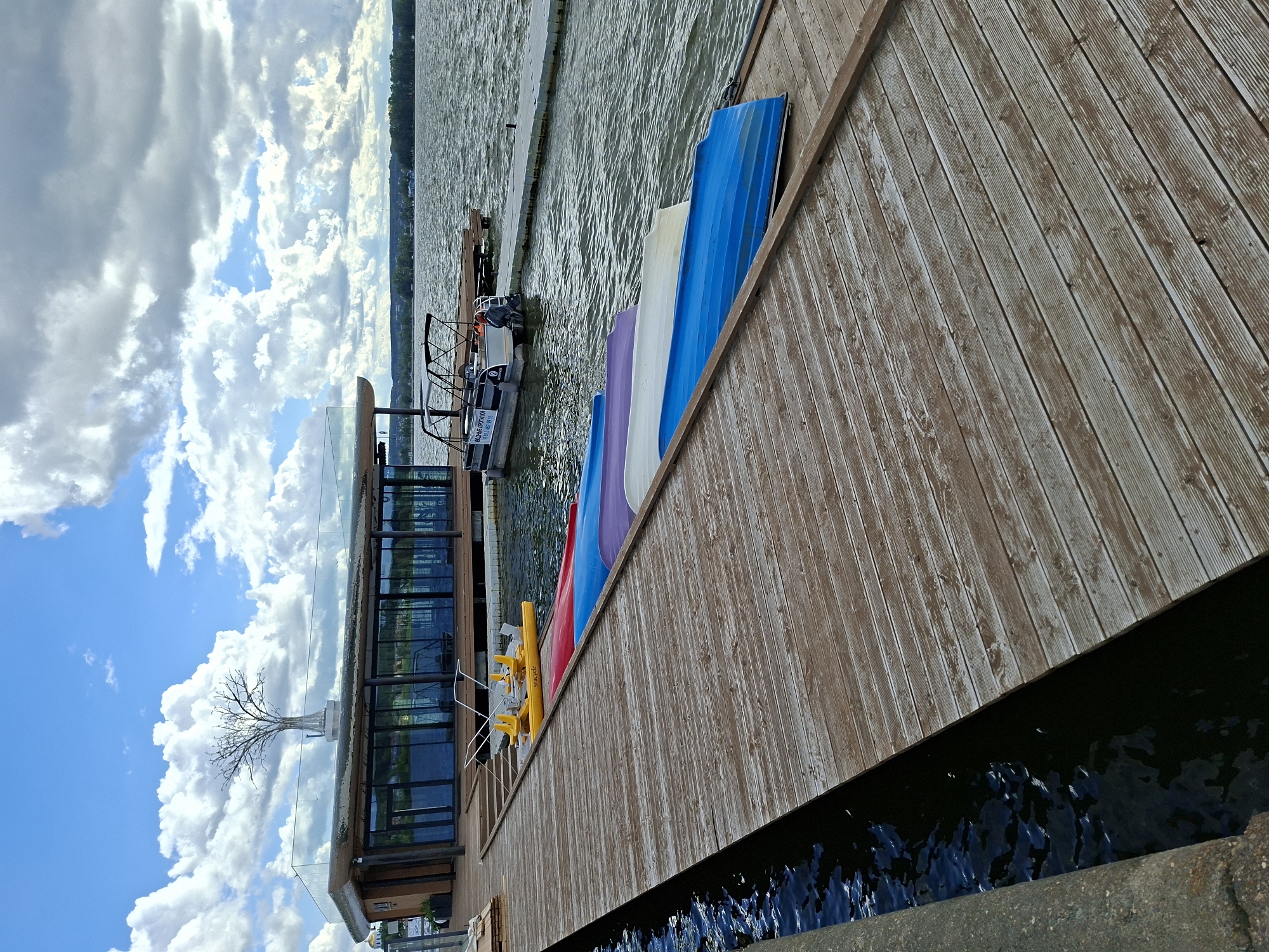 The image shows a wooden dock on a lake in Votkinsk, Russia. On the dock, there are several kayaks stacked, a yellow pedal boat, and a small motorboat tied to a floating dock. In the background, there is a modern building with large glass windows, topped with a decorative tree sculpture. The lake is calm, with ripples on the surface, and the sky is partly cloudy. The overall scene depicts a waterfront leisure area.