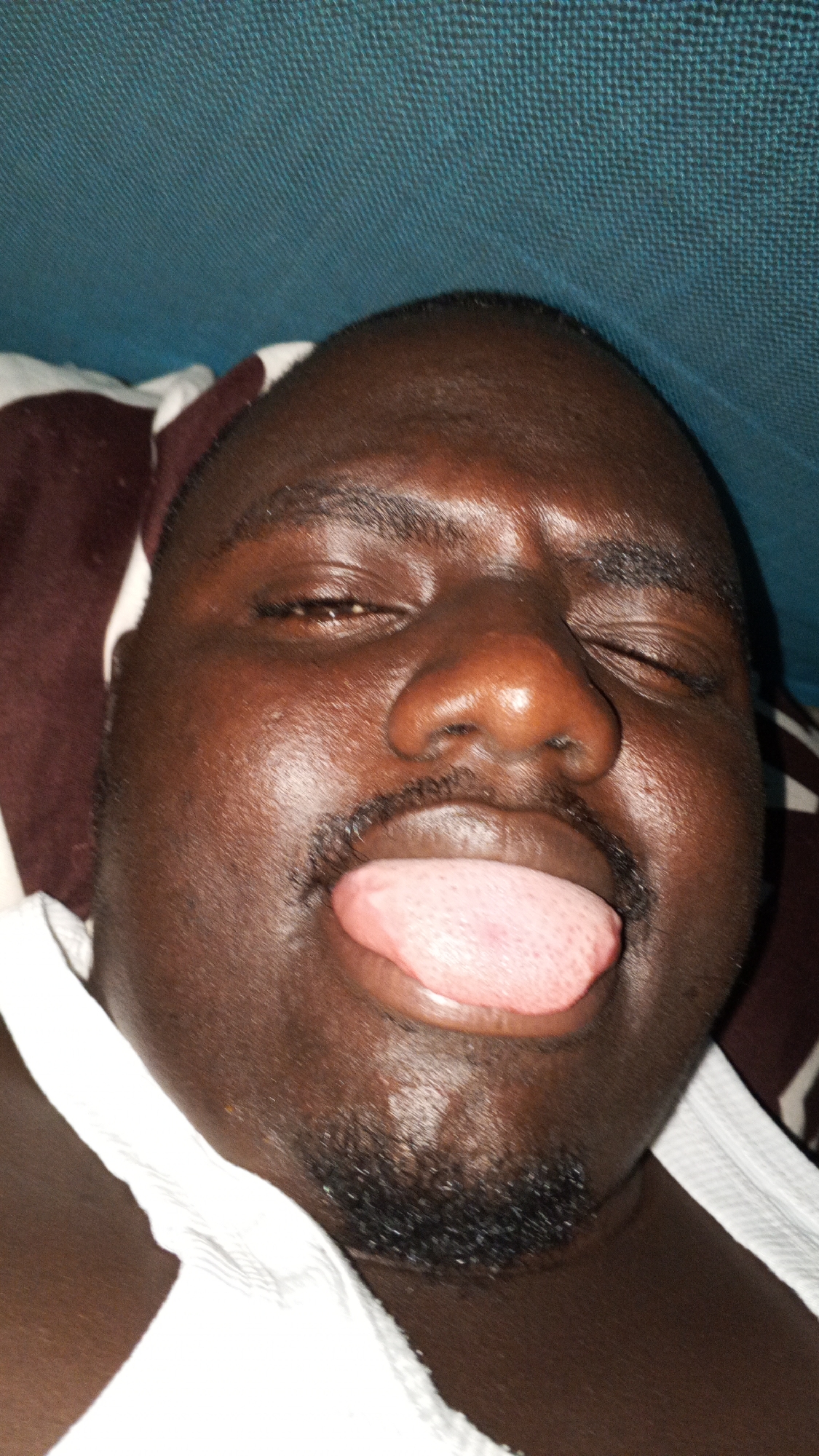 A close-up photograph shows a dark-skinned man with a mustache and beard, lying down with his eyes mostly closed and tongue sticking out. He is wearing a white garment. A brown and white patterned pillow is behind his head, and a textured blue surface is visible above him. The location is unknown.