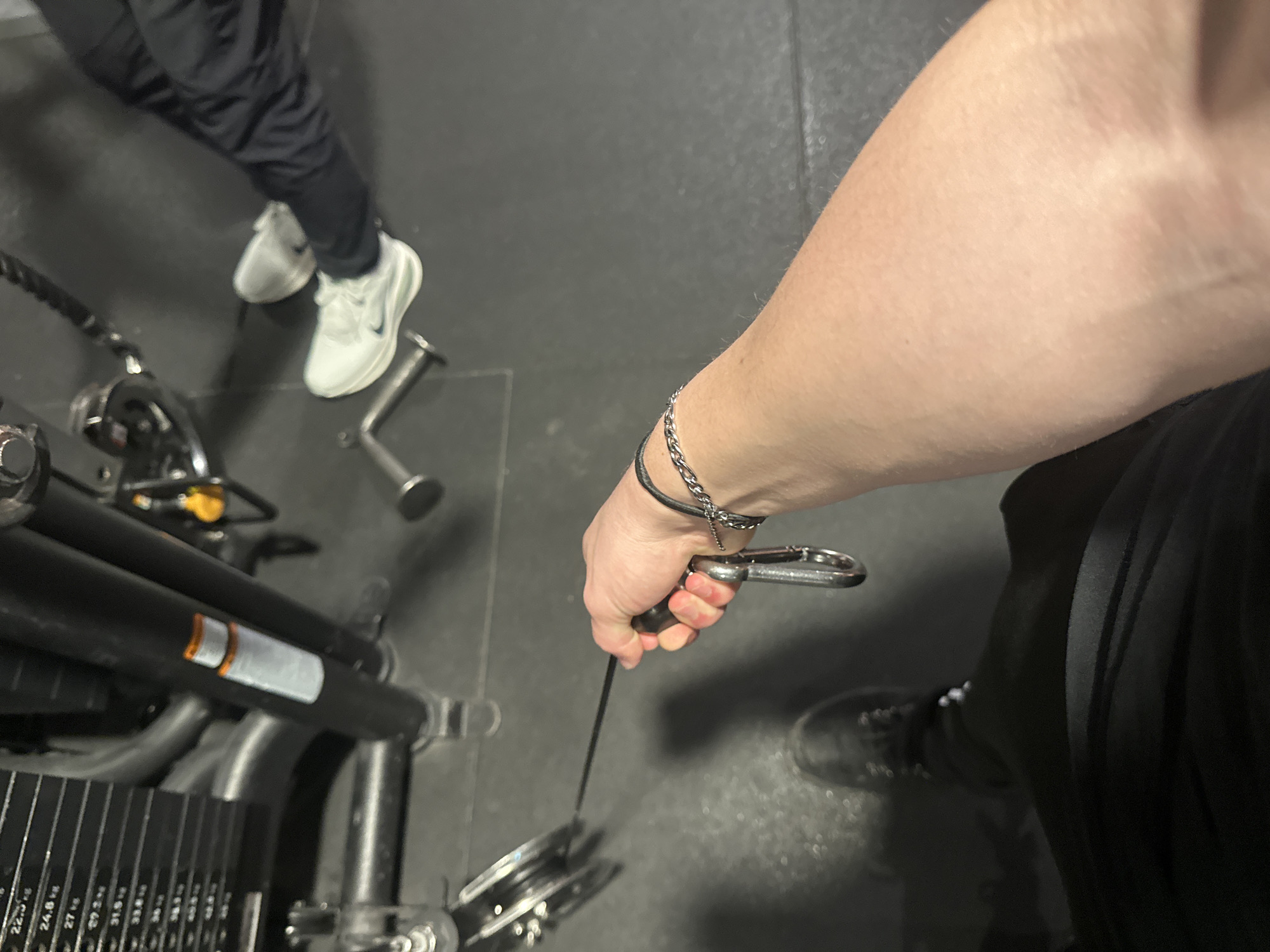 The image displays an indoor scene, consistent with a gym environment, potentially located in Schagen, Netherlands, though the specific address within the city cannot be determined from the visual information. The perspective is an overhead, downward-angled shot.
In the foreground, a fair-skinned right arm extends from the lower left toward the center, with the hand gripping a black handle. A silver-colored metal carabiner connects the handle to a thin black cable that extends toward a pulley system on the mid-left. A black band and a silver-colored chain bracelet are worn on the right wrist. Partially visible on the lower left is the primary subject's lower torso, clad in dark athletic wear, and a dark shoe.
To the upper left, a black cable machine is visible, featuring a weight stack with multiple silver plates. Visible weight increments include 22.5 kg, 24.8 kg, 27 kg, 31.5 kg, 33.8 kg, 36 kg, 38.3 kg, 40.5 kg, 42.8 kg, 45 kg, 47.2 kg, and 49.5 kg. An orange and white warning label is affixed to one of the machine's vertical black support columns.
In the background, on the right side of the frame, the lower legs and feet of a second individual are visible. This person is wearing dark trousers and white athletic shoes with dark soles, consistent with the Nike brand.
The floor is a dark gray or black material, likely rubber matting, showing faint seam lines. Two small, T-shaped metal floor attachments or handles are present on the floor. The lighting appears to be artificial, illuminating the interior space.