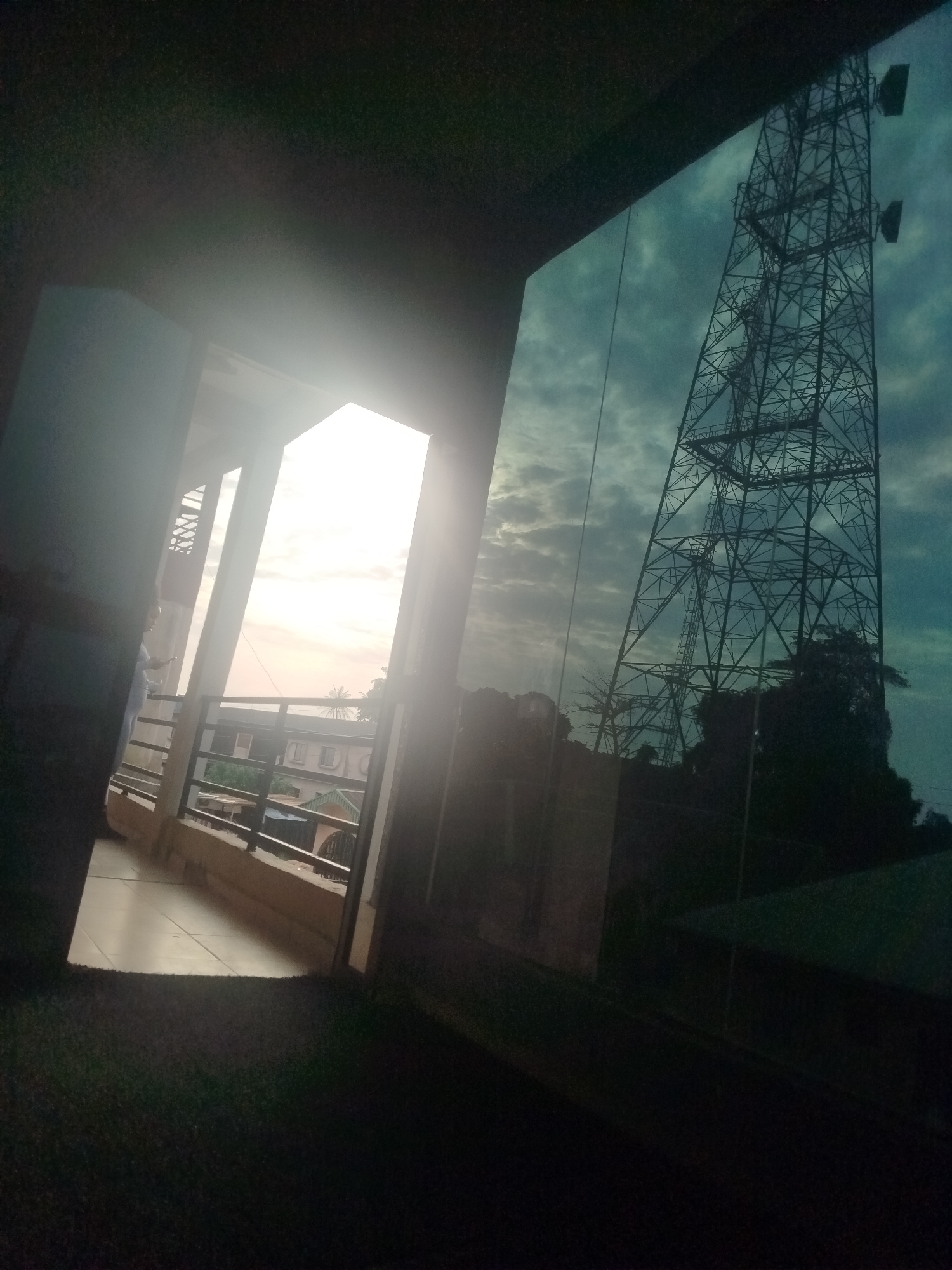The image shows a view from inside a building looking out through a large window and an open doorway. On the right, the window reflects a tall, metallic telecommunications tower against a cloudy, turquoise sky. The tower is complex and latticed, with several platforms and antennas visible. In the distance, silhouetted trees and other structures are partially visible.
To the left, through the open doorway and a balcony or veranda, a bright, overcast sky with a soft sunrise or sunset casts a warm glow. The balcony has metal railings and a tiled floor. Beyond the balcony, residential buildings with multiple stories and pitched roofs are visible. A person, possibly a woman, is standing on the balcony, holding a mobile phone. The foreground is dark, with a textured surface that appears to be carpet or a similar fabric. The overall lighting suggests either early morning or late afternoon.