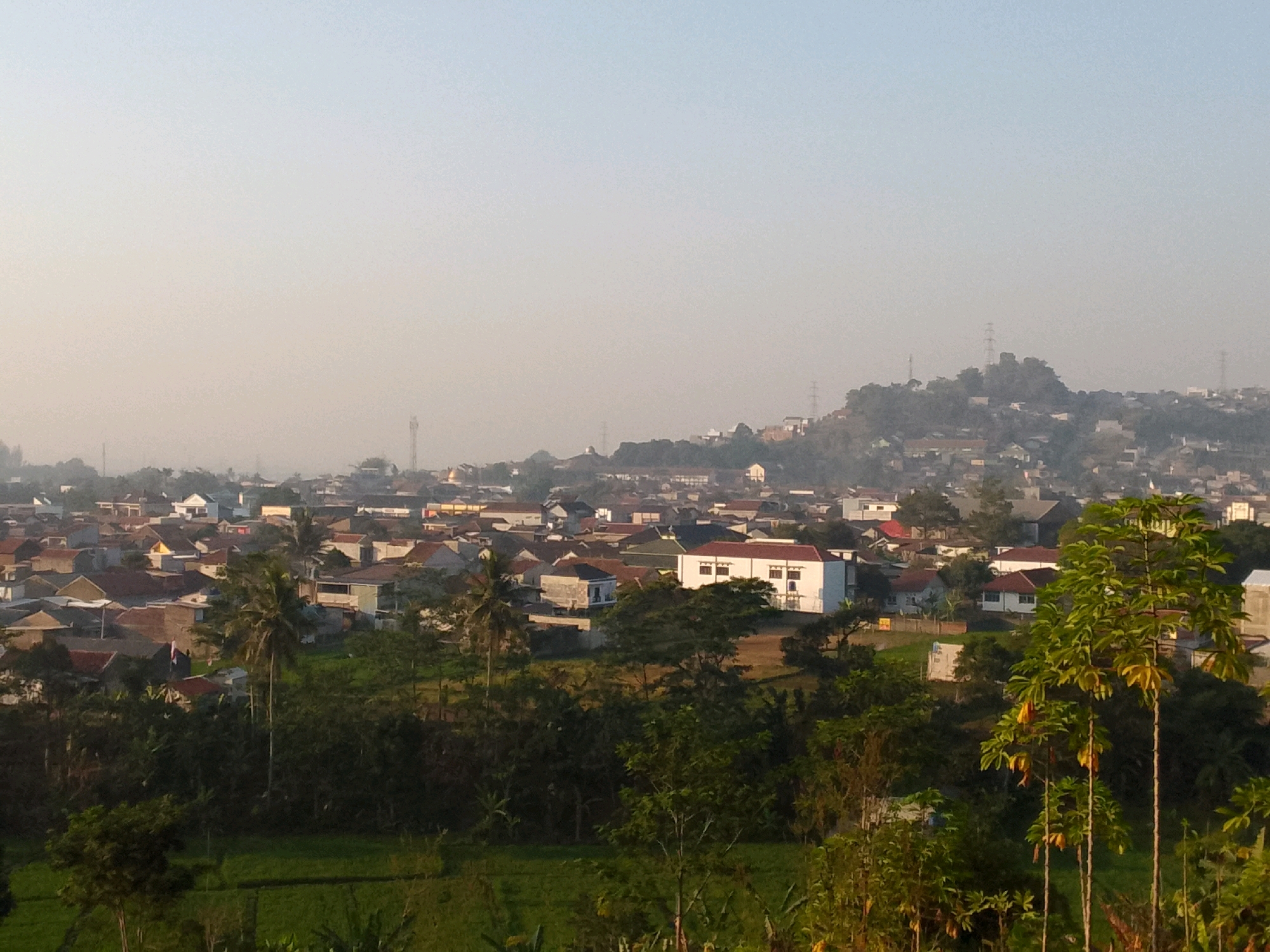 The image displays a panoramic view of a residential area in Padalarang, Indonesia, likely captured during early morning or late afternoon due to the soft, hazy lighting. The scene is dominated by a densely packed neighborhood with numerous houses characterized by red-tiled roofs and white or light-colored walls. A prominent white building with dark-paned windows stands out in the midground.
In the foreground, lush green vegetation and trees, including palm trees, are visible, suggesting a tropical or subtropical climate. The terrain appears to be hilly, with houses built on slopes and the landscape extending towards a distant, hazy horizon. Several telecommunication towers are discernible on the elevated areas in the background, indicating the presence of infrastructure within or near the settlement. The overall impression is one of a suburban or semi-urban environment with a mix of residential buildings and natural greenery.
