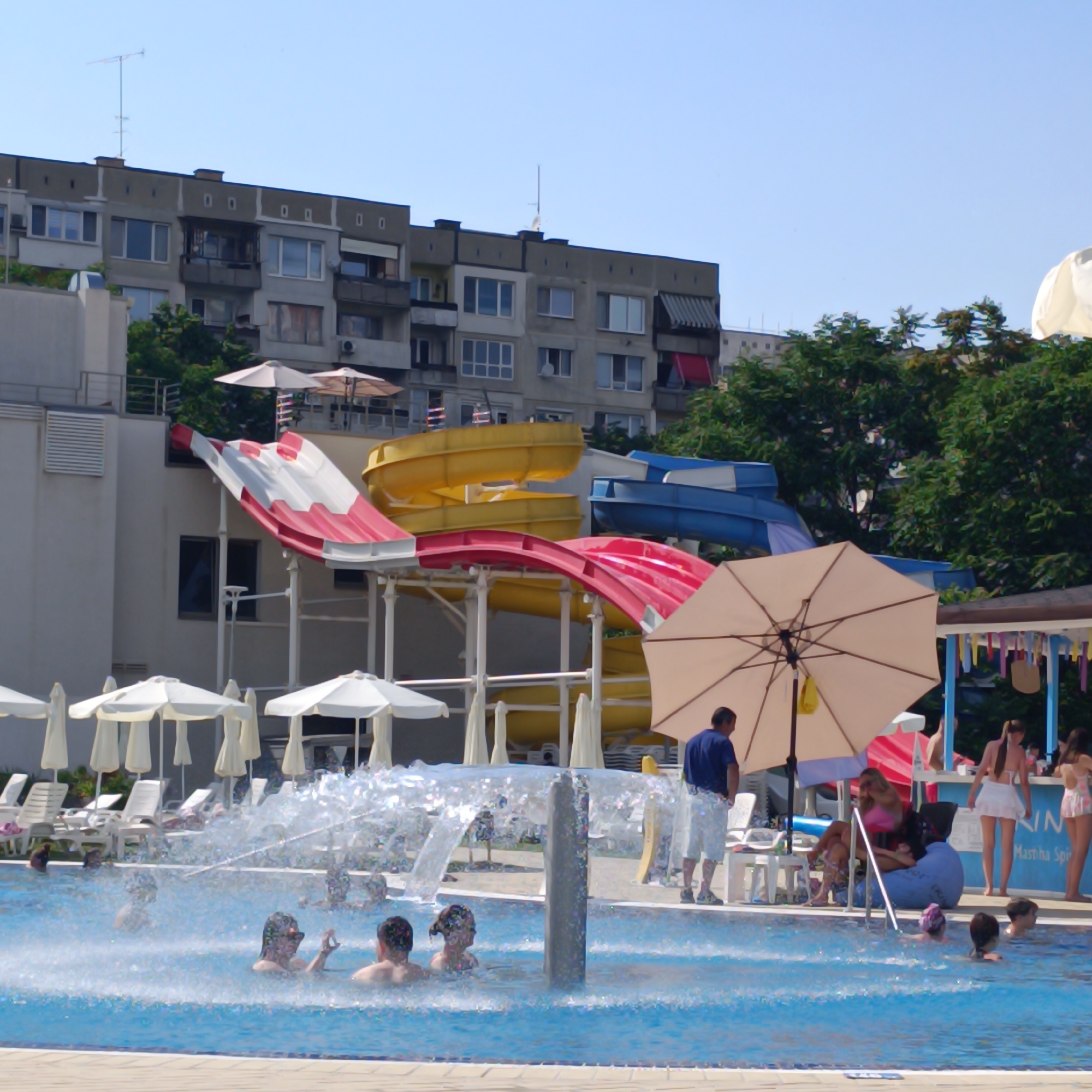The image depicts a sunny day at an outdoor water park in Sofia, Bulgaria. Several individuals are enjoying the pool, with some splashing in the water near a fountain. In the background, a large water slide complex with red, white, and yellow sections is visible, along with a blue water slide. The setting includes rows of white lounge chairs and umbrellas, suggesting a leisure environment.
Dominating the upper portion of the image are apartment buildings, characteristic of urban Bulgarian architecture. Green trees provide a natural contrast to the man-made structures. The sky is clear blue, indicating good weather.
One notable detail is a person in a blue shirt and white shorts standing near a large, beige umbrella, seemingly observing the pool area. In the background, near the water slides, a bar area is set up with people attending to it. Some of the visible text includes "IN" and "Mastiha Spi". The overall atmosphere appears relaxed and recreational, with people enjoying a summer day at the water park.