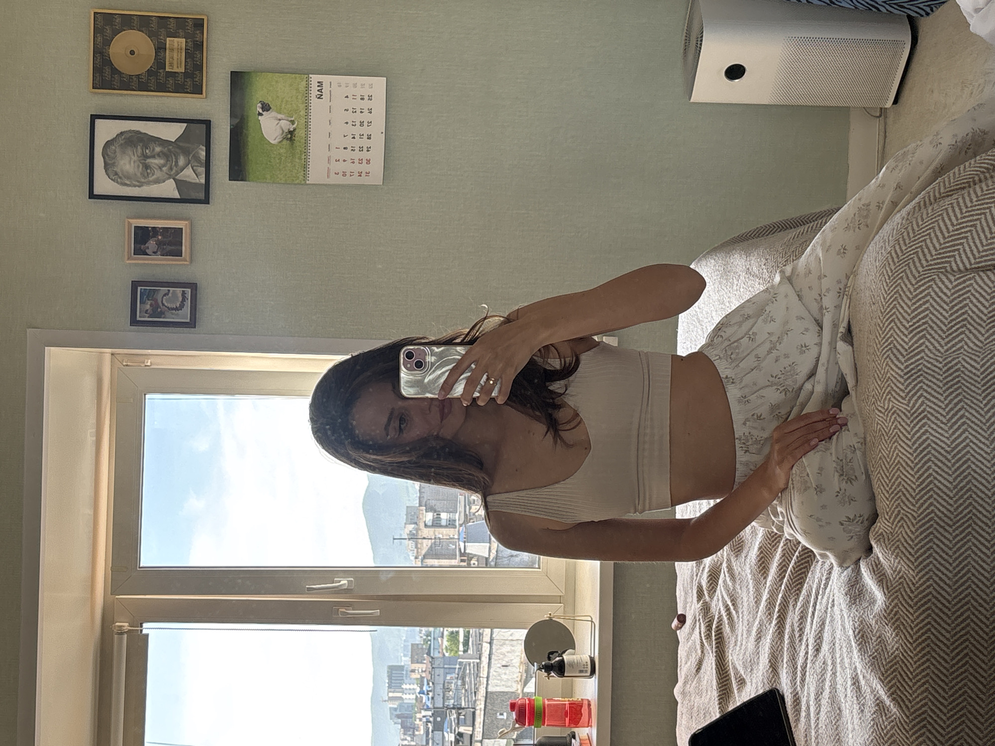 A young woman captures a mirror selfie in a well-lit room, possibly a bedroom, overlooking a city. Her face is partially obscured by the smartphone she holds, reflecting her brown hair and a light-colored top. She stands beside a bed adorned with a light beige chevron-patterned blanket and a floral-patterned pillow. A dark tablet lies on the bed.
The room features light green, textured wallpaper. On the wall, several items are displayed: a framed gold-colored disc with repetitive "Aaaaa" text, a black and white portrait of an older man, and two smaller framed photographs beneath it. A calendar to the right shows a white dog on a green lawn, with "NAM" at the top and clearly visible days of the week and dates from 1 to 31. A white air purifier is visible near the ceiling.
A large window offers a view of a city with multiple buildings and what appear to be distant mountains under a clear, bright blue sky, indicating daytime, likely morning or early afternoon. On the windowsill, two water bottles (one red, one green), a small round mirror, and a black bottle are arranged. The setting suggests a casual, lived-in apartment in an urban environment, consistent with Krasnoyarsk, Russia.