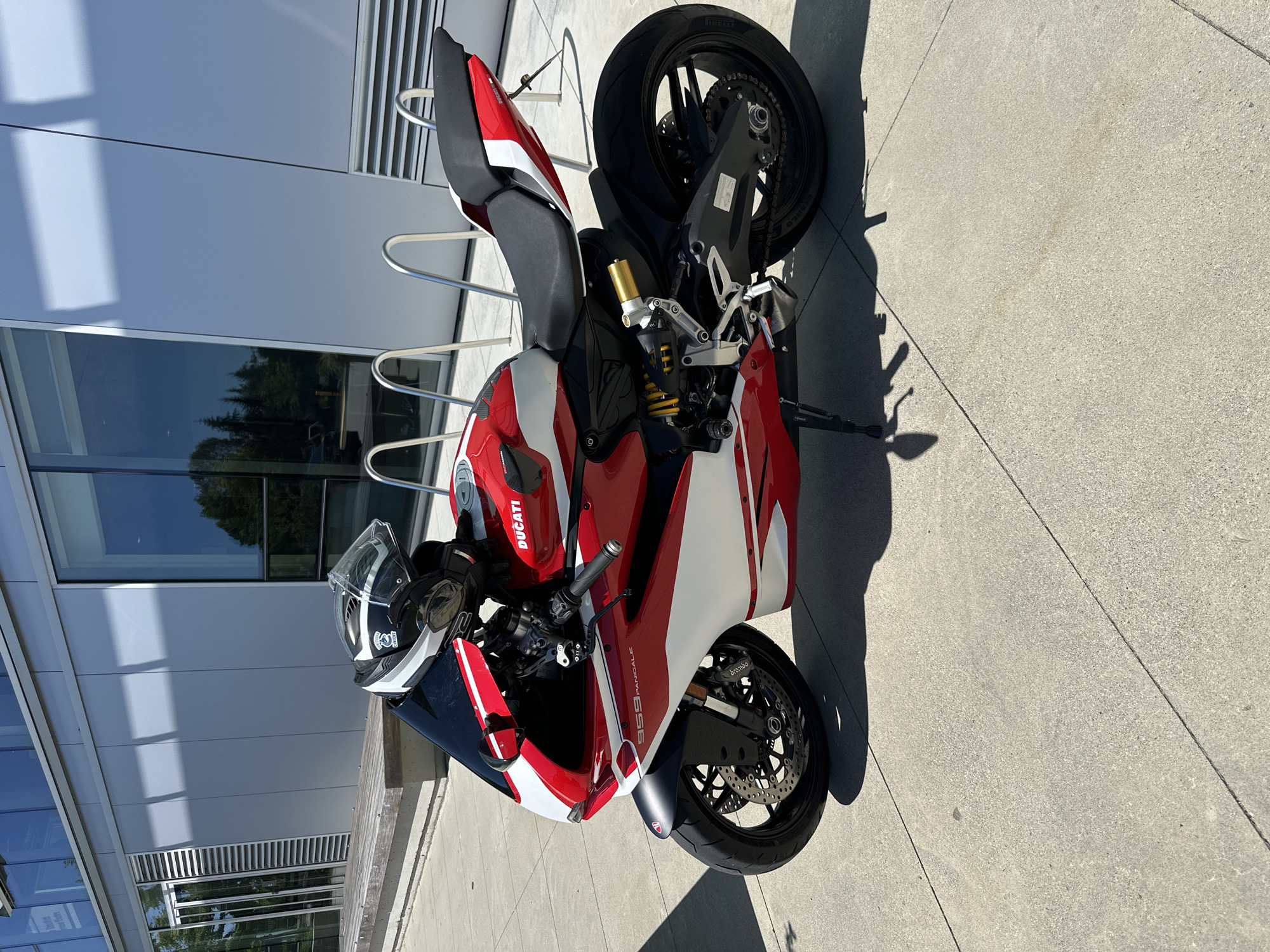 A red and white Ducati motorcycle is parked on a concrete sidewalk in front of a modern building with large windows. A black helmet with white accents is placed on the seat of the motorcycle. The building appears to be an office or institutional building with a silver facade and horizontal paneling. There is a wooden bench in the foreground, and a bicycle rack behind the motorcycle. The scene is brightly lit by sunlight, casting shadows on the ground. The location is identified as Burnaby, Canada.