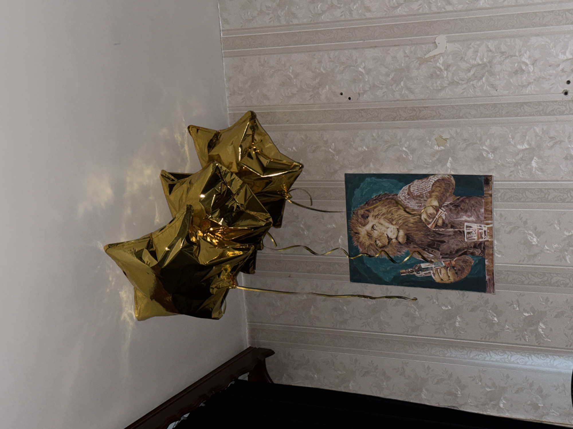 The image shows a room with a bed, gold star-shaped balloons, and a framed picture hanging on the wall. The wallpaper is patterned with a floral design and horizontal stripes. There are imperfections on the wallpaper, such as tears and patches. The picture on the wall depicts a lion holding a weapon. The bed frame is dark brown, and the bedding is black. The location is Khimki, Russia.