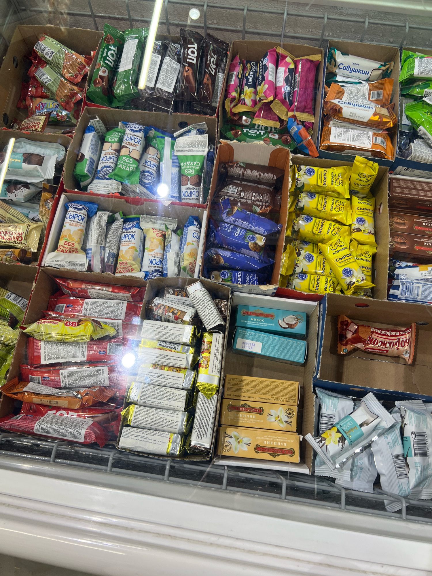 The image shows an indoor view of a freezer display case filled with various packaged food items, likely in a grocery store or market. The display is divided into several compartments containing products like ice cream bars, chocolate bars, and snack packs. The items are neatly arranged in cardboard boxes and individually wrapped. The lighting suggests the presence of overhead fluorescent lights, reflecting off the glass of the freezer. There are no people visible in the image. The overall environment appears to be clean and organized, typical of a retail food establishment. The location is identified as Osh City, Kyrgyzstan.