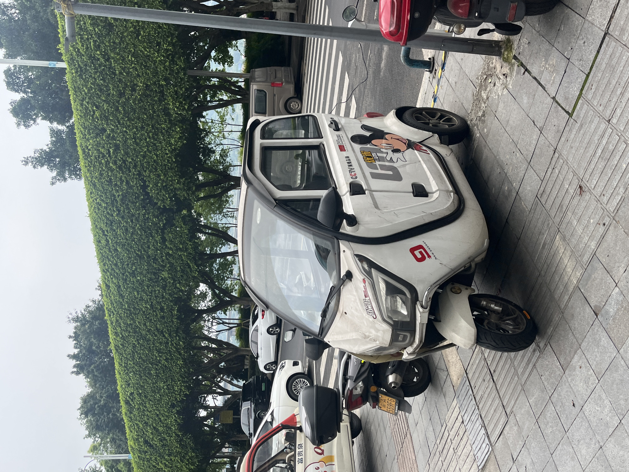 The image depicts a white electric tricycle with a small cabin, parked on a tiled sidewalk next to a hedge-lined street. The tricycle features a Mickey Mouse sticker on its side. The scene is in Jiangjin, China.
