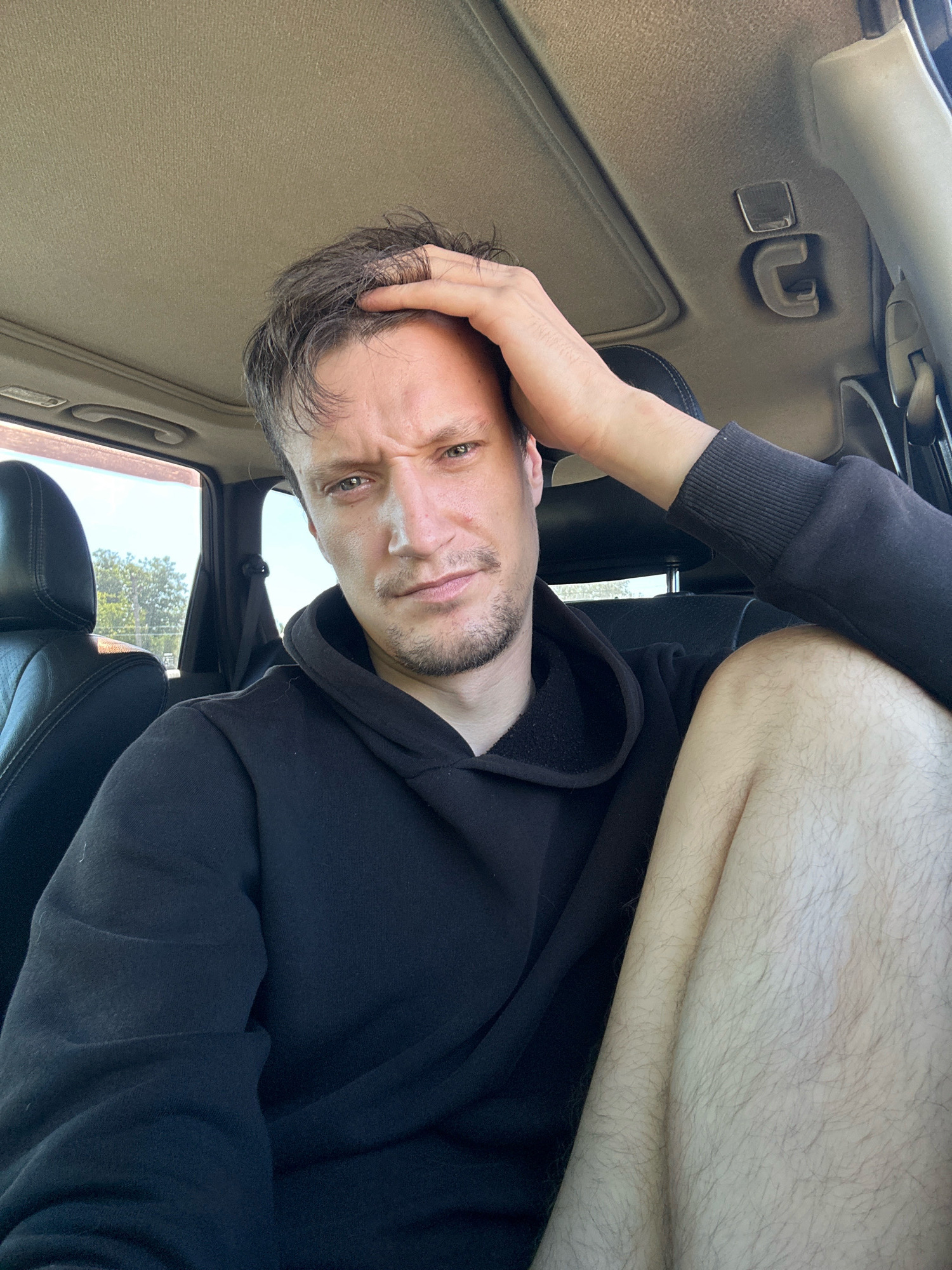 A medium shot shows a man in a black hoodie sitting in the driver's seat of a car. He is looking at the camera with his hand on his head, his eyebrows furrowed. The man has light skin and dark hair, with stubble on his chin and upper lip. His left leg is visible, and it appears to be hairy. The car's interior is visible, with black leather seats and a beige headliner. The sun is shining outside, and trees and a fence can be seen through the car window. The man's expression suggests he may be stressed or tired. The image appears to have been taken during the day.