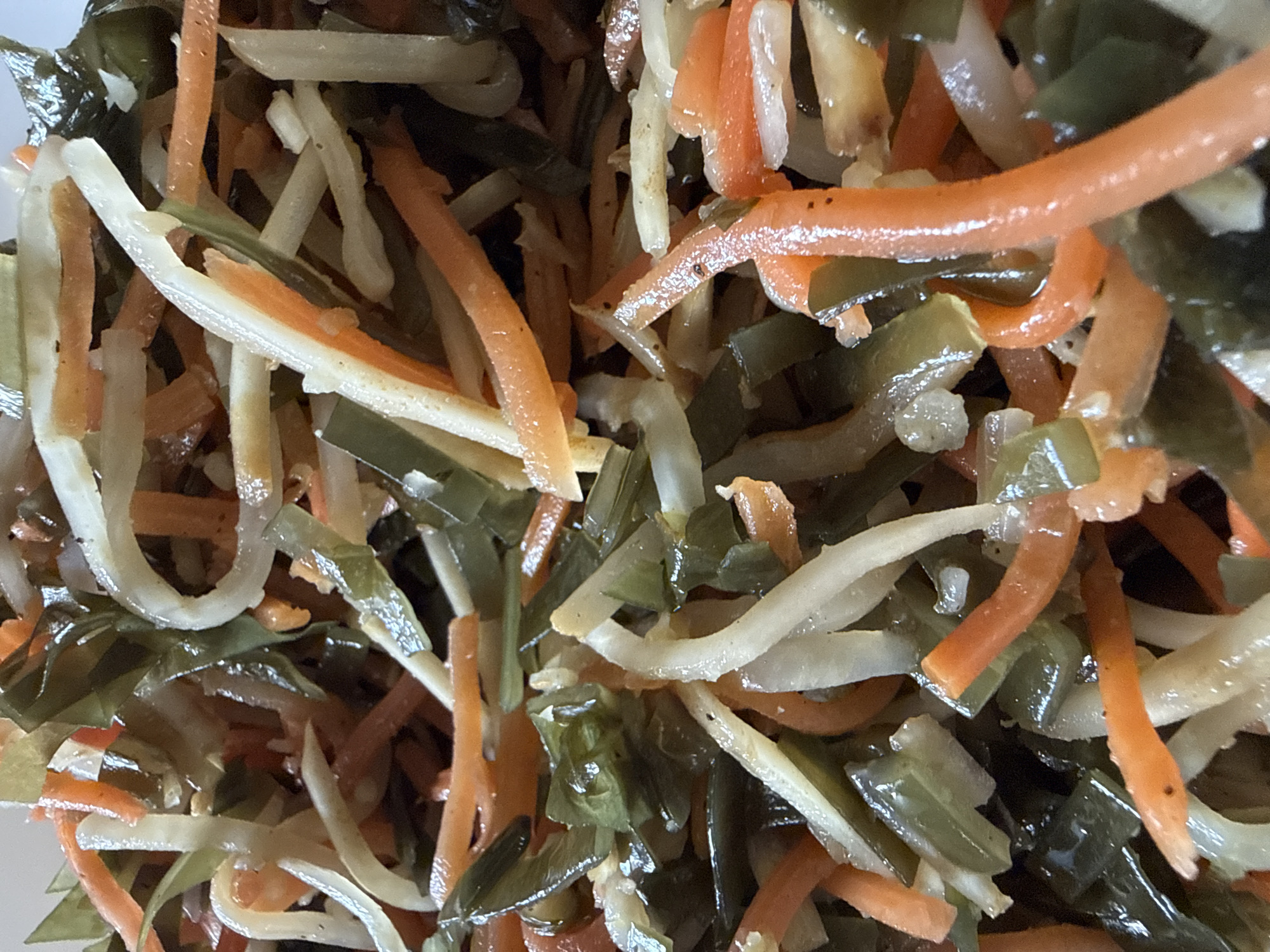 The image is a close-up, overhead view of a salad. The salad contains julienned vegetables in shades of orange, white, and dark green. The orange pieces appear to be carrots, and the white pieces resemble daikon radish or jicama. The dark green pieces are likely seaweed, such as wakame or kelp. The vegetables are mixed together and appear to be dressed in a light, glossy sauce. There are no people visible in the image. The background is out of focus and appears to be the interior of a bowl or container. There is no information in the image to indicate a location in Okhtyrka, Ukraine.