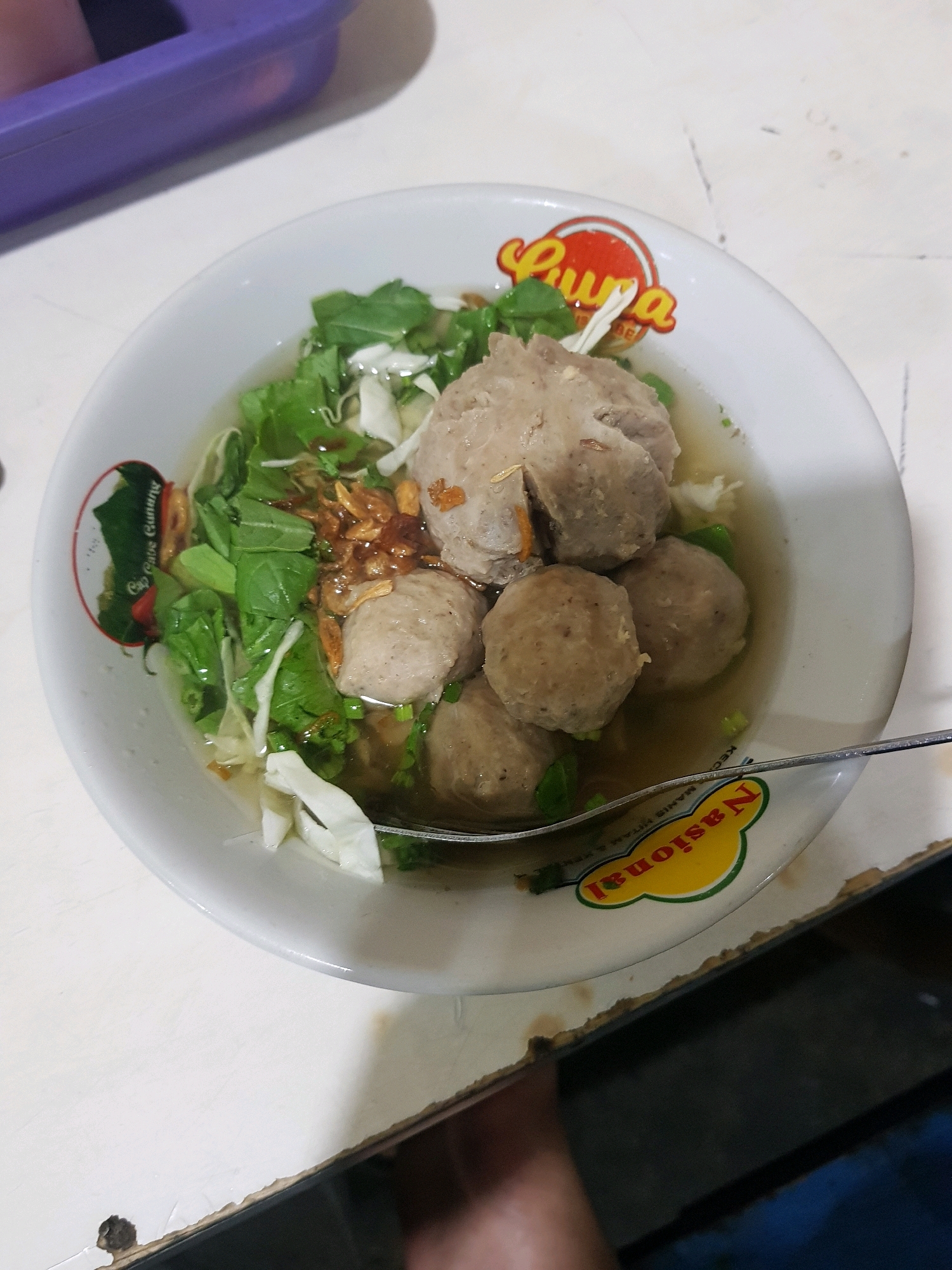 A close-up, eye-level shot shows a bowl of bakso, a popular Indonesian meatball soup, on a white speckled table. The bowl is white with a red and yellow logo on the rim, depicting a red chili pepper with the text "Cap Garpu Gunung" and a yellow oval with "Nasional" in red text. The bowl contains several large, round, light-brown meatballs, submerged in a clear broth. Green leafy vegetables, possibly bok choy, and shredded white cabbage are visible, along with fried shallots. A silver spoon rests on the rim of the bowl. In the background, a blurry purple object is visible on the left, and the table surface has some brown stains and scuff marks. The scene appears to be in a casual dining setting.