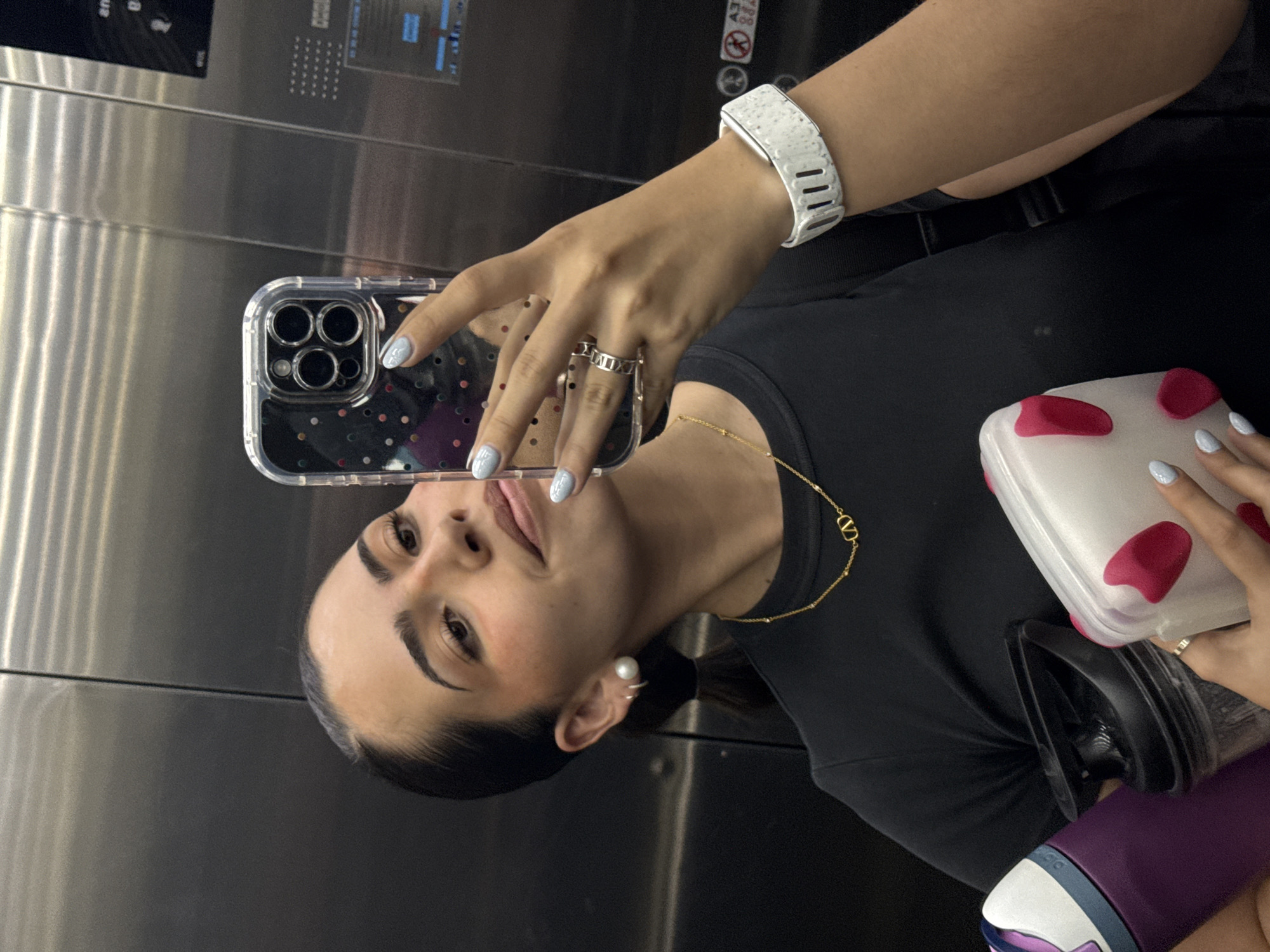 A woman is taking a selfie in an elevator. She is holding a smartphone with a clear case in her right hand, and a lunchbox and water bottle in her left. The elevator interior is stainless steel. The woman is wearing a black top, a white smart watch, rings on her fingers, a gold necklace and a pearl earring. The location is within the urban region of San Bernardino Tlaxcalancingo, Mexico, as elevators are predominantly found in multi-story buildings and are not common in rural areas.