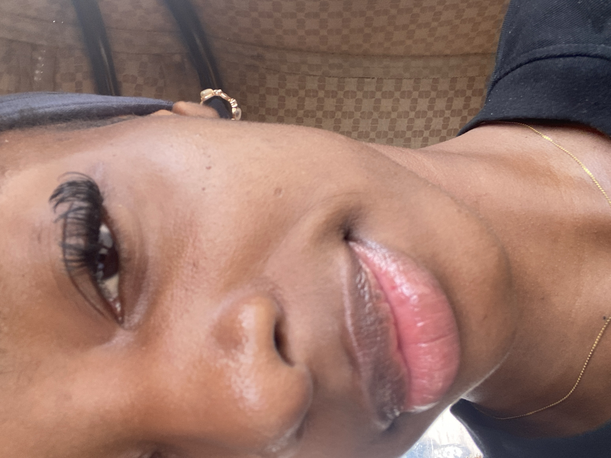 The image is a close-up, frontal portrait of a young Black woman, likely in her late teens or early twenties. She has dark skin, long, dark eyelashes, and full lips with a glossy sheen. She is wearing a dark headband and a dark shirt, with a delicate gold chain visible around her neck. A gold hoop earring with small stones is also visible on her right ear. The background appears to be an indoor setting with a patterned wall, possibly made of fabric or wallpaper, in shades of brown and beige. The lighting is soft and appears to be natural light coming from the front, illuminating her face and creating subtle highlights on her skin and lips. The overall impression is one of a casual, personal photograph. The provided information about the location in Jos, Nigeria, is not visually verifiable from the image itself.