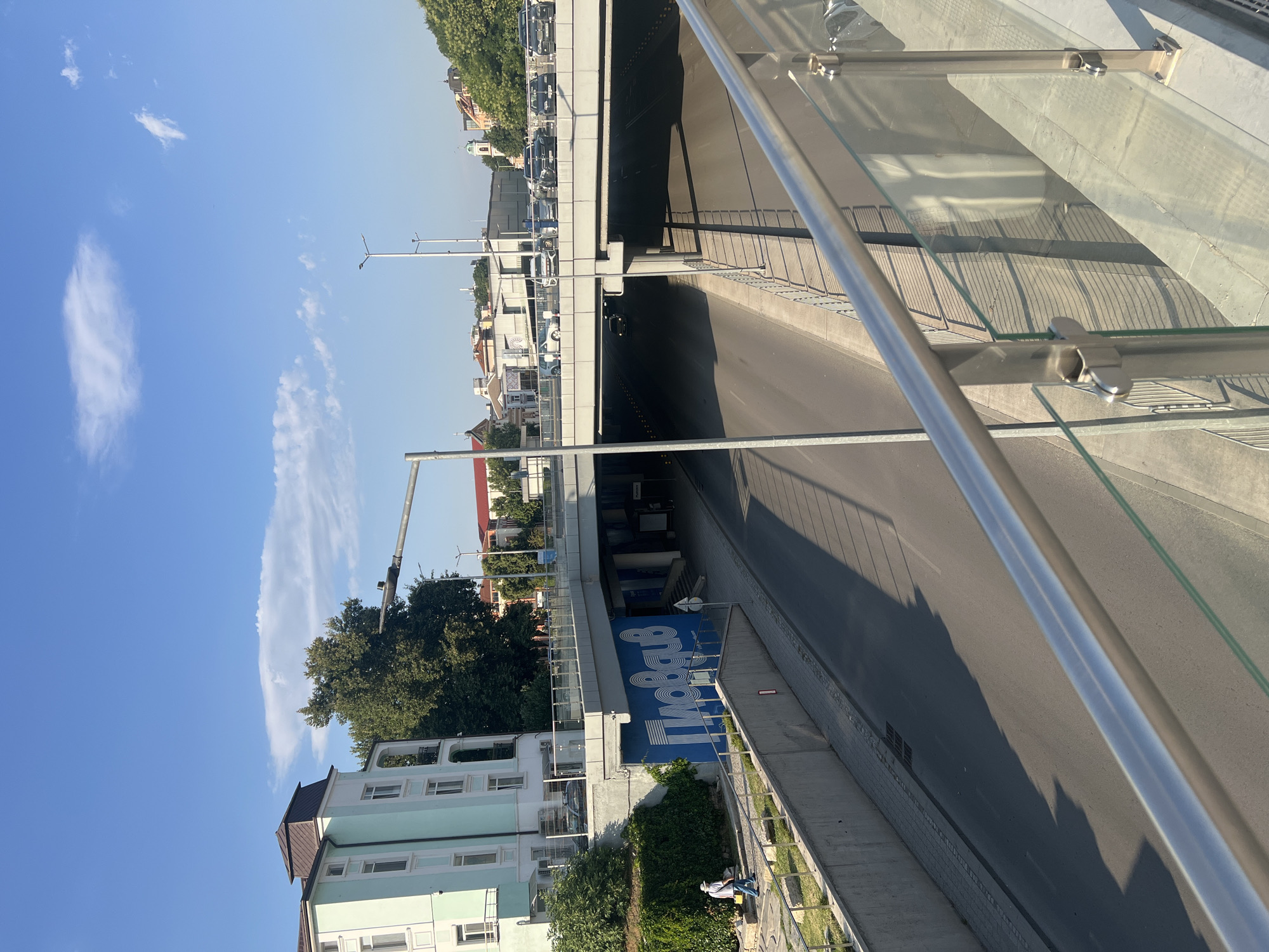 The image captures a view of a street-level underpass and an overpass in the city of Plovdiv, Bulgaria. The underpass is a wide, paved roadway with vehicles visible in the distance, partially obscured by shadows. The overpass, constructed with concrete and railings, features cars parked along its edge. On the left side of the image, a light blue building with white trim and multiple windows stands tall, bordered by a lush green hedge. Below the overpass and next to the blue building, a large blue mural with white text is prominently displayed. A person in a light-colored shirt and hat is walking on a paved path in the foreground, near the hedge. The sky above is clear blue with a few scattered clouds, indicating a daytime scene. The overall environment suggests an urban setting with significant infrastructure.