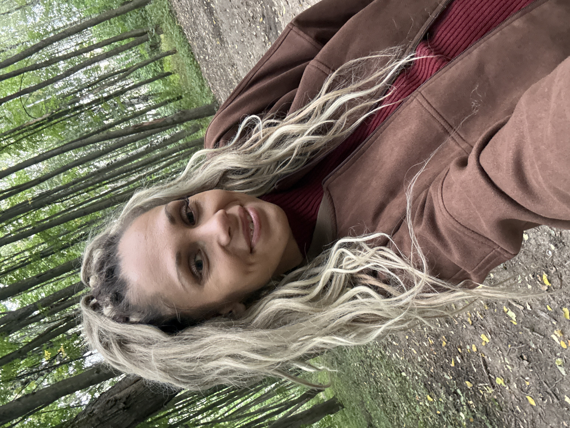 A woman with blonde hair styled in braids and a ponytail smiles at the camera. She is wearing a maroon turtleneck sweater and a brown zip-up jacket. She is outdoors in a forest with tall trees and fallen leaves on the ground. The lighting suggests it is daytime.