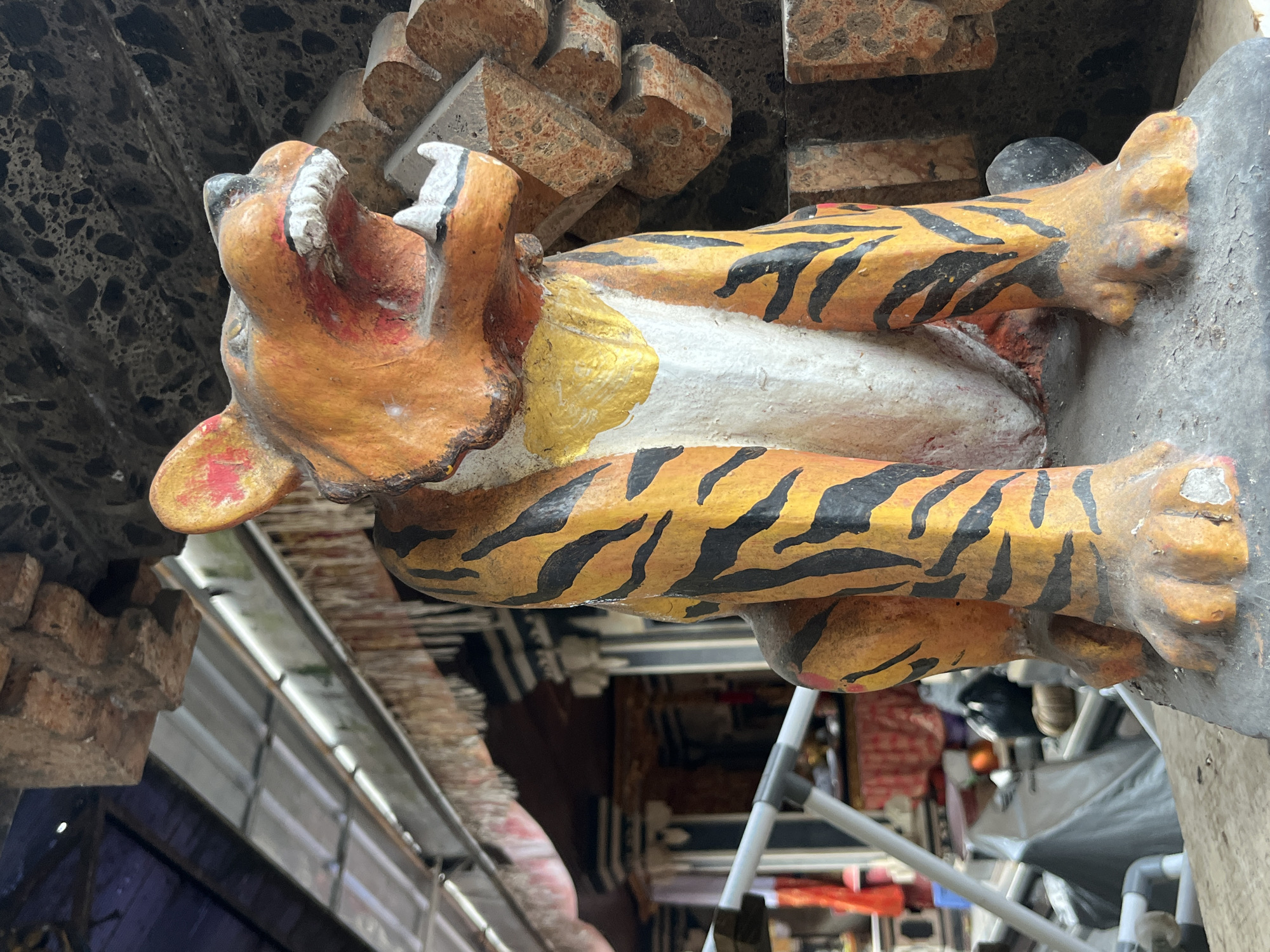 An outdoor, eye-level, close-up shot shows a brightly colored, weathered statue of a tiger in Kuta Selatan, Indonesia. The tiger is depicted in an aggressive pose, with its mouth open, revealing sharp white teeth and a red interior. Its body is painted with vivid orange and black stripes, and a yellow chest area. The statue is positioned on a stone base, with a rustic, textured background of orange and gray stone. In the background, a covered structure with a corrugated metal roof and a dark blue tarp is visible. Frayed, decorative fabric hangs from the edge of the structure. The area appears to be an outdoor marketplace or temple, with glimpses of colorful textiles, metal scaffolding, and various objects on shelves. The overall impression is one of vibrant, cultural artistry set against a utilitarian, outdoor environment.