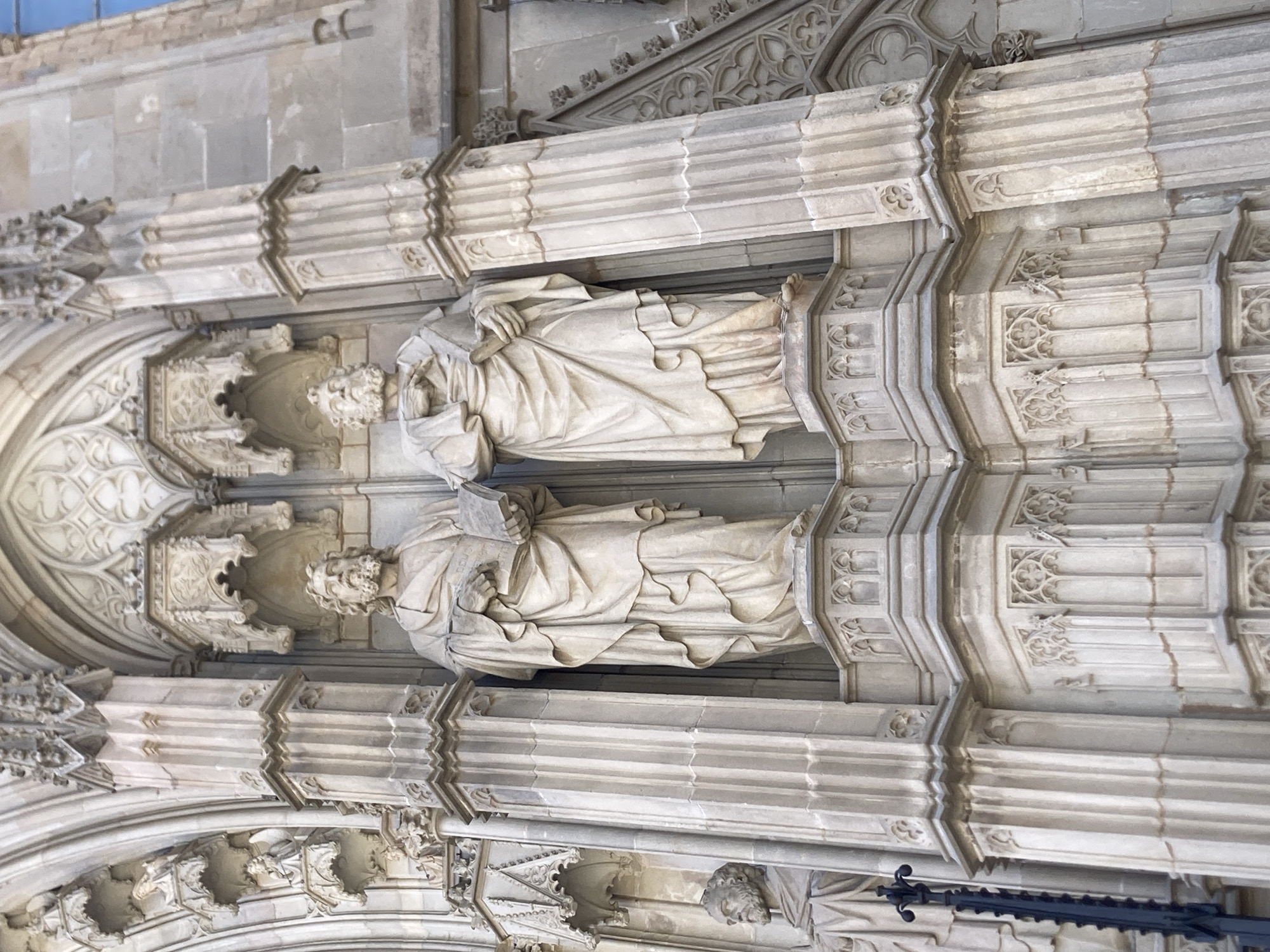 This is a close-up, low-angle, exterior shot of Gothic architecture, likely a cathedral or similar historical building in Barcelona, Spain. The image is vertically oriented and focuses on intricate stone carvings and sculptures.
The primary subjects are two life-sized stone sculptures of robed figures, likely saints or historical personages, embedded within niches of the building's facade. Both figures are depicted holding what appear to be books or tablets. The upper sculpture is more fully visible, showing a bearded face and hands delicately holding an open book. The lower sculpture is partially obscured but also appears to be holding a book. The figures are set against a backdrop of ornate architectural details, including pointed arches, ribbed vaults, and decorative tracery. The stone appears weathered, suggesting the age of the structure.
The scene is static, showcasing the static nature of the sculptures and the building. There is no discernible activity or event taking place, other than the implied passage of time indicated by the weathered stone. The lighting is bright and diffused, suggesting it is daytime, though the exact time cannot be determined. The weather appears clear, as there are no visible signs of rain or clouds. The architectural style, particularly the pointed arches and the detailed carvings, strongly points to a Gothic origin, and the provided location context of Barcelona, Spain, suggests it is likely a famous landmark there. No visible text is present in the image.