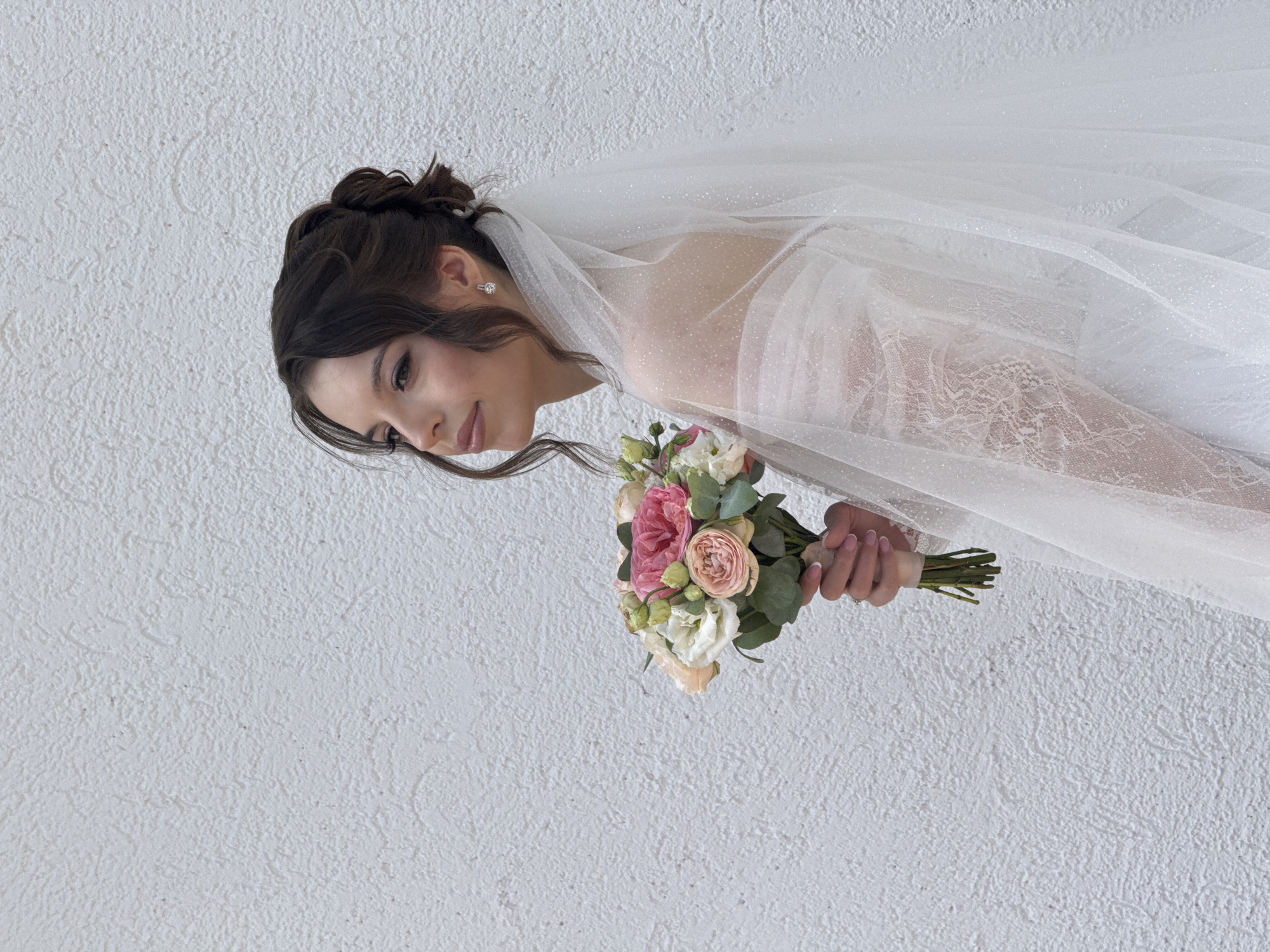 The image shows a bride in a white wedding dress and veil, holding a bouquet of flowers. She is looking down and to the side with a soft smile on her face. Her hair is styled in an updo, and she is wearing diamond earrings. The bouquet contains roses in shades of pink and white, along with greenery. The bride is standing in front of a textured white wall. The scene appears to be indoors, with soft lighting that suggests it could be daytime. The image evokes a feeling of joy and anticipation, fitting for a wedding. There is no visible text in the image.