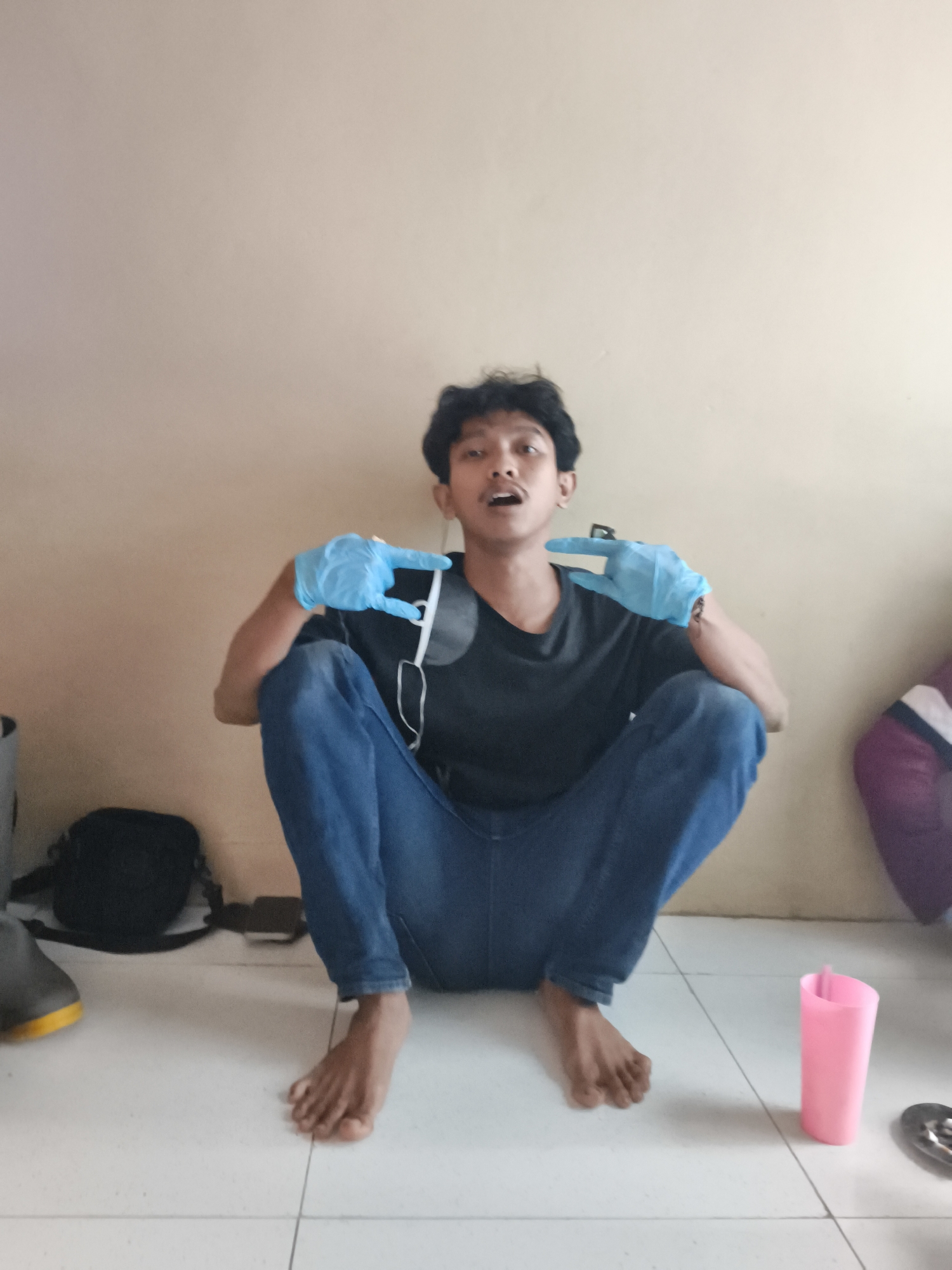 A young adult male with curly black hair and light brown skin is crouched on a tiled floor, facing the camera. He is wearing a dark t-shirt, blue jeans, and light blue disposable gloves. His mouth is open, and his hands are raised in front of him, with the gloved index fingers pointing towards each other. A white cord, possibly from headphones, hangs from his neck. Behind him, a beige wall serves as the backdrop. To his left, a black bag and a pair of grey and yellow boots are visible on the floor. To his right, a pink plastic cup stands near a circular metallic object. The overall impression is a casual indoor setting, likely a home or a common area, potentially in Pekalongan, Indonesia, given the provided context.