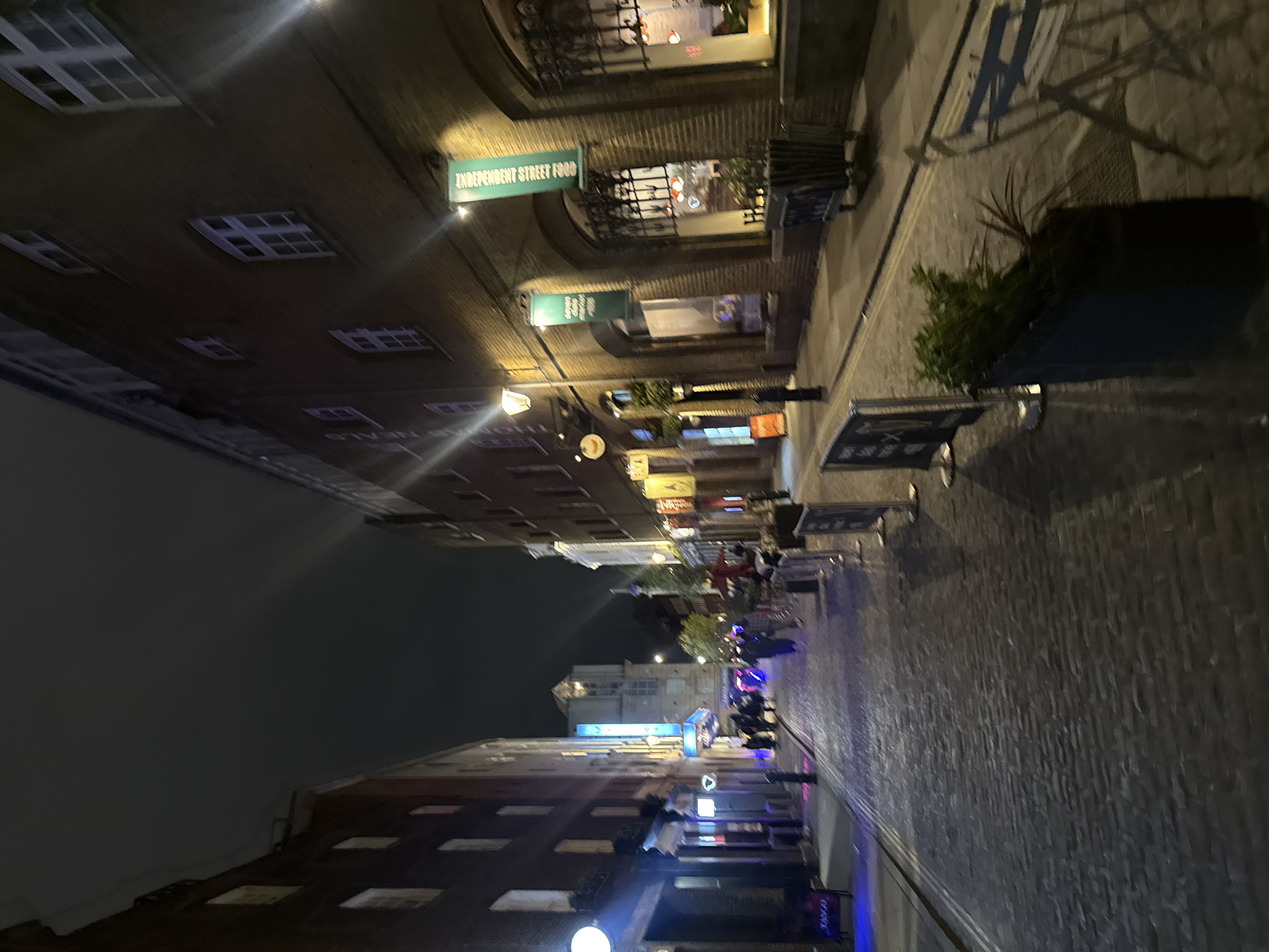 The image shows a street in London, United Kingdom at night. The street is lined with buildings, mostly brick, with several stores and restaurants. There are signs hanging from the buildings, one of which says "Independent Street Food". People can be seen walking down the street, and there are some tables and chairs set up outside the buildings. The street is lit by streetlights, and the sky is dark. The pavement is cobbled.