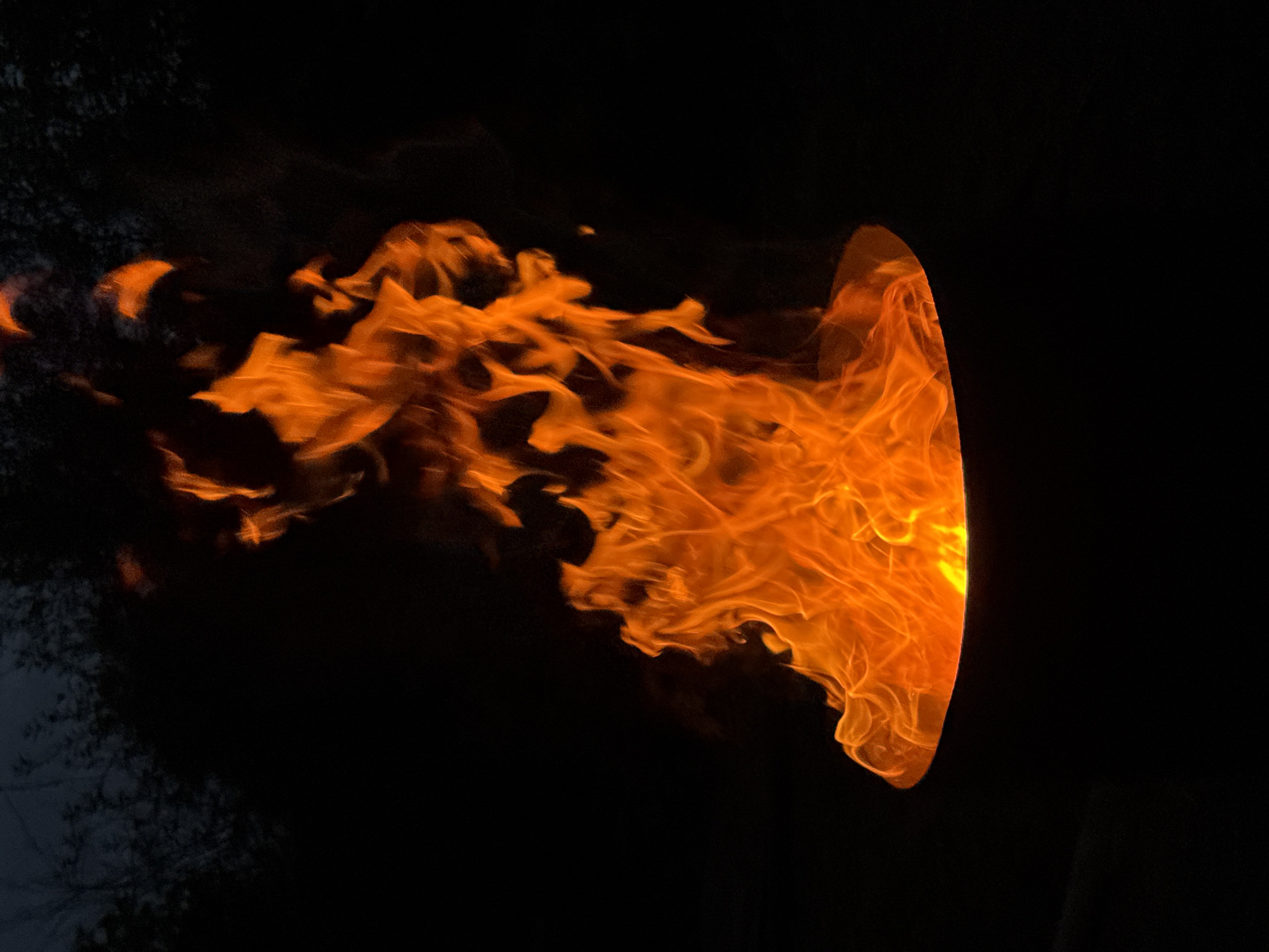 The image depicts a close-up view of a fire burning intensely in what appears to be a metal container, possibly a fire pit or a cauldron. The flames are predominantly orange and yellow, with wisps of white at the hottest points. The background is entirely black, suggesting a nighttime setting, with the faint silhouette of dark trees visible at the very top of the frame. The scene is dark and atmospheric. There are no people visible in the image. The location in Chaikovsky, Russia, is not discernible from the visual content of the image itself.