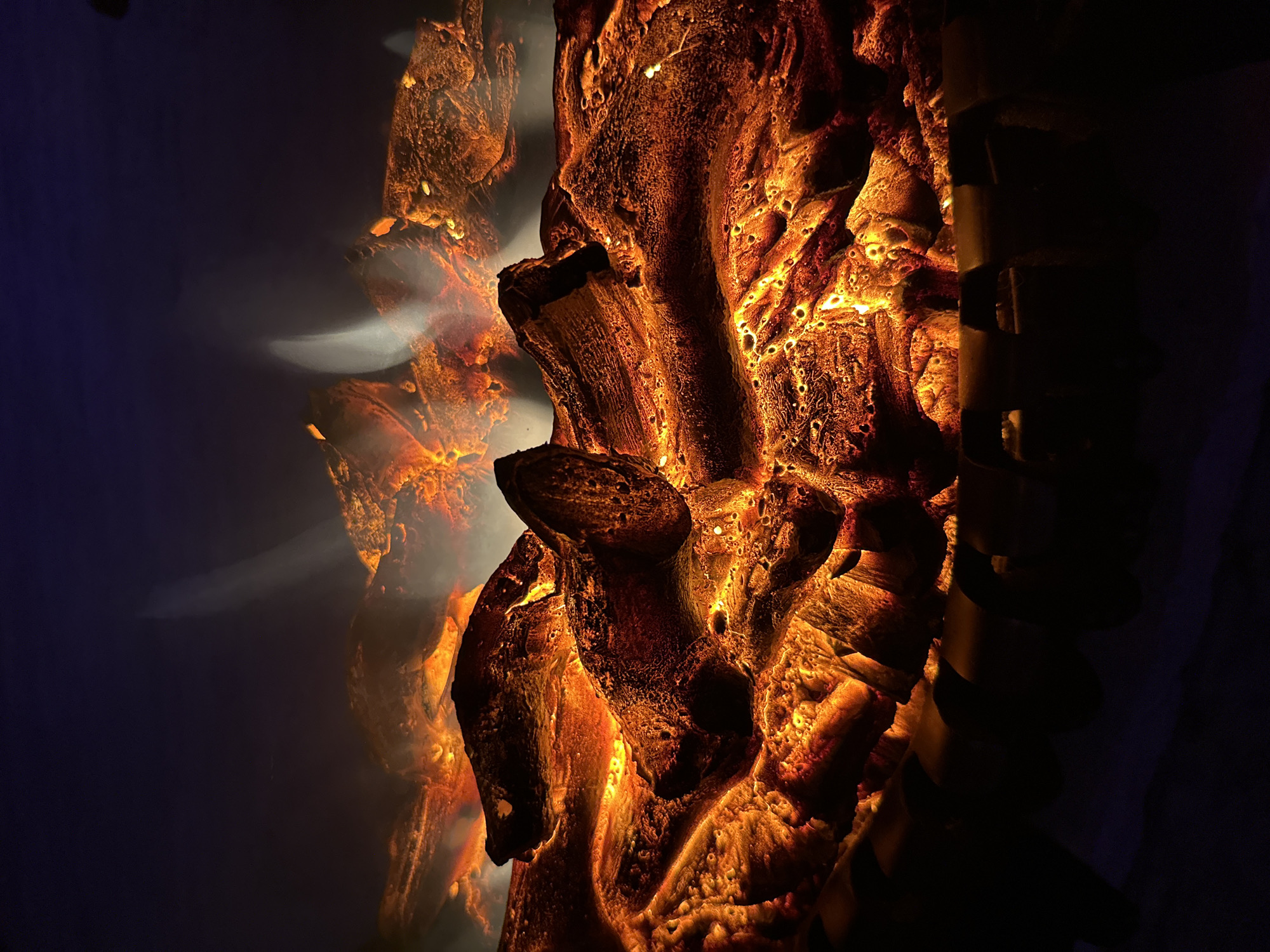 A close-up, low-angle shot captures the simulated glow of an electric fireplace. The scene is dominated by textured, orange and yellow light emanating from a pile of dark, log-like structures. These logs appear to be made of a material that mimics burnt wood, with crevices and irregular surfaces catching the light. Wisps of white light, suggesting flames or smoke, rise into the dark, shadowy background. The bottom of the image shows a row of dark, curved metallic elements, possibly part of the fireplace's housing or a grate. The overall impression is one of artificial warmth and light against a dark, indistinct environment.