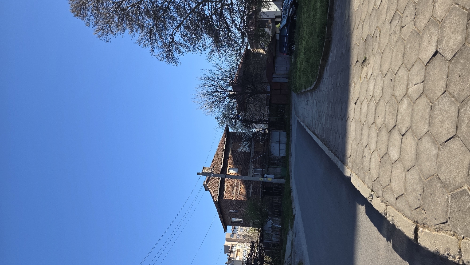 The image captures a street scene in Blagoevgrad, Bulgaria. A two-story brick building with a reddish-brown tiled roof stands prominently on the left side of the frame. It appears old and somewhat dilapidated, with visible wear and tear on the facade. A utility pole is situated directly in front of the building, with power lines stretching across the sky. To the left of the building, there is a clear blue sky with some bare tree branches visible in the upper corner. A paved sidewalk constructed with hexagonal concrete blocks runs along the right edge of the image. A narrow asphalt road is visible between the building and the sidewalk. The scene suggests a residential area, possibly on the outskirts of the city. The lighting indicates it is a sunny day. There are no people visible in the frame.