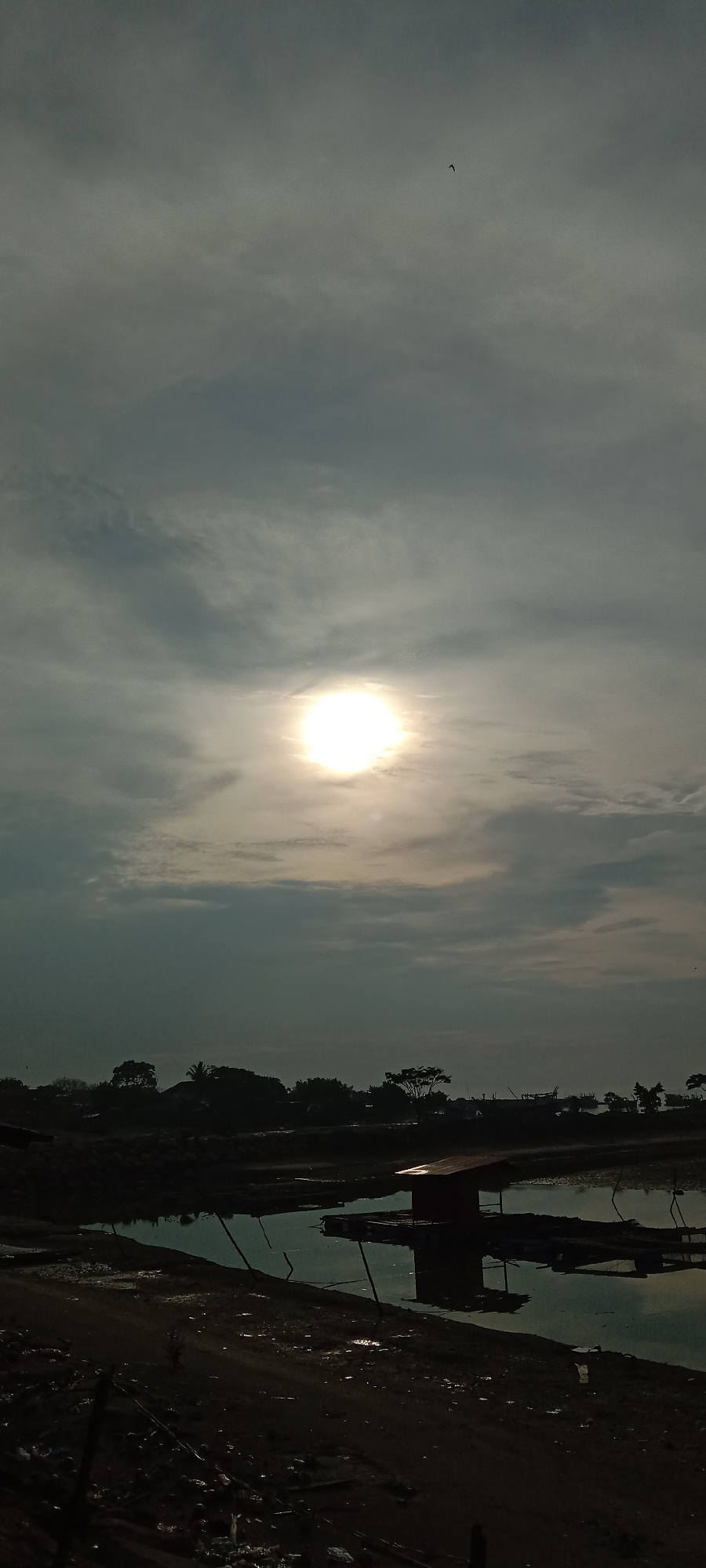 The image shows a sun in a cloudy sky over a landscape. In the foreground, there is a body of water, with a small structure in it. The landscape in the middle ground includes trees and possibly other vegetation. There is a bird visible in the sky. The general location is likely Lhokseumawe, Indonesia.