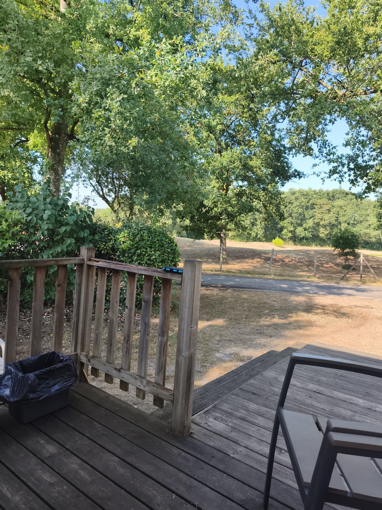 The image depicts a wooden deck with a railing on the left side, overlooking a sun-drenched landscape. A black trash bag is visible on the deck, placed in a dark-colored bin. In the foreground, a portion of an outdoor chair is present. Beyond the deck, a gravel path leads into a dry, grassy field dotted with trees. In the distance, a dense forest borders the field, under a clear blue sky. The scene appears to be an outdoor seating area, likely in a park or campsite, given the rustic wooden structures and the natural surroundings. The lighting suggests it is daytime, with the sun casting shadows.