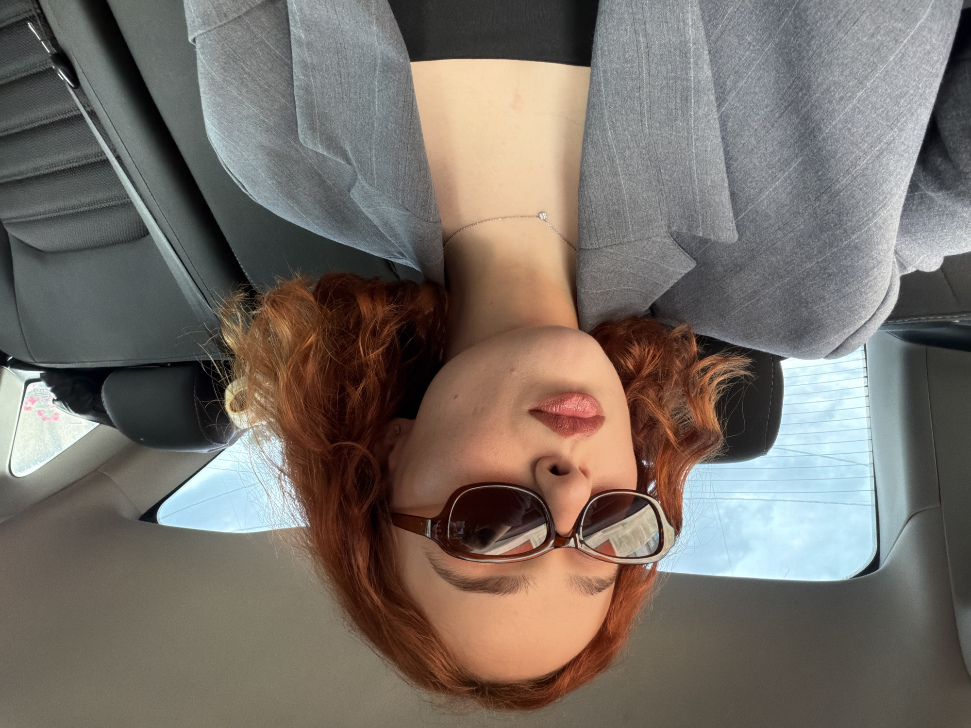A woman with auburn curly hair and brown sunglasses is visible from the neck up. She is wearing a gray pinstripe blazer over a black top and a thin silver necklace with a small pendant is visible at her neck. The photograph appears to be taken inside a car, based on the interior elements like the seat, headrest, and window visible in the background. The sky is visible through the rear window. There is no evidence to determine that the location is in the city of Tbilisi, Georgia.