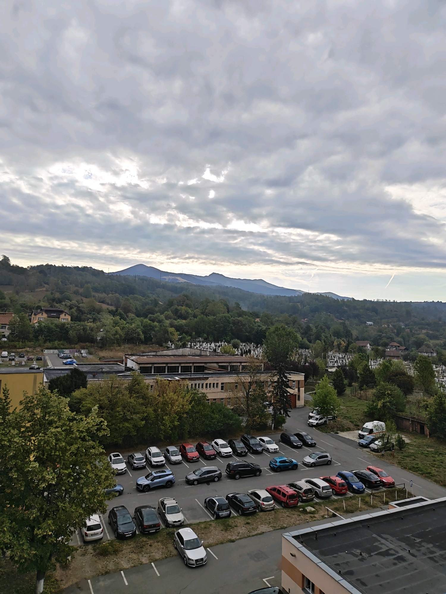 A high-angle, full shot captures a parking lot filled with cars, situated near a cemetery and a forested hillside under a cloudy sky. The cars are parked in rows, with a variety of makes and models visible, including sedans, SUVs, and a white van. The cemetery is to the right of the parking lot, marked by rows of white headstones. The background features a dense forest covering a hilly landscape, with mountains visible in the distance. A few buildings, including what appears to be a school or hospital with a yellow facade, are also present. The lighting suggests it is daytime, possibly overcast. The image appears to be taken in Petroșani, Romania, given the context. There are no people visible in the image.