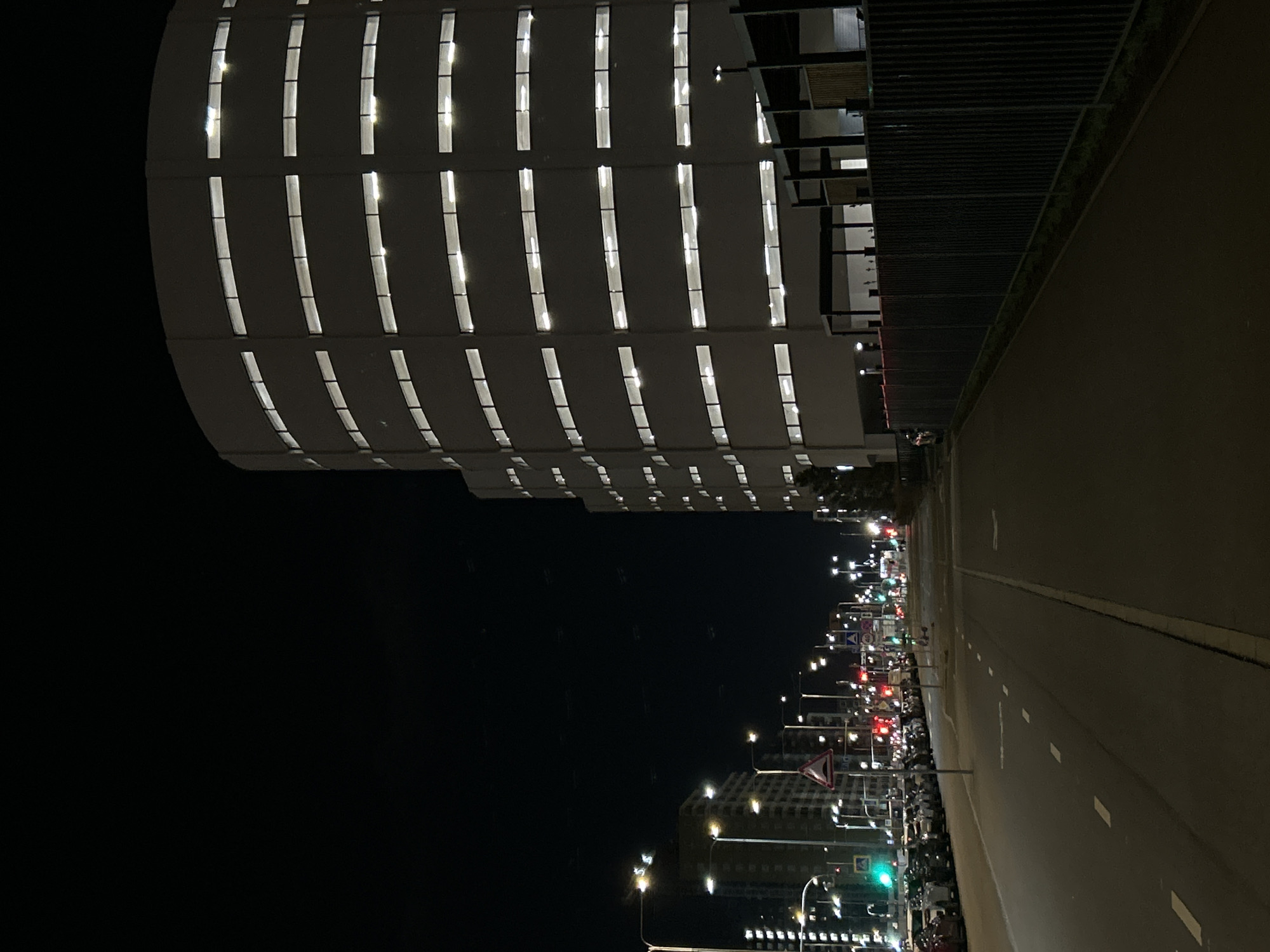 This media file depicts an urban street scene at night in Saint Petersburg, Russia, characterized by artificial light illuminating modern architecture.
On the left, a large, multi-story building with a distinctly curved facade dominates the view. Its exterior is a uniform dark grey, punctuated by numerous vertically elongated windows that glow brightly with white light, casting rectangular shafts into the darkness. These windows are arranged in a regular, staggered pattern across multiple floors, suggesting a contemporary design, possibly a parking structure or a large commercial building. Adjacent to this building, and forming a boundary with the road, is a darker section featuring horizontal slats or a metal grate, possibly part of a fence or another architectural element.
To the right, a wide, empty street extends into the background, clearly delineated by white dashed lane markings. The road is flanked by a series of glowing streetlights that stretch into the distance. Further along the street, a cluster of urban infrastructure becomes visible, including additional buildings with illuminated windows, traffic lights displaying both red and green signals, and various road signs. One triangular red and white road sign, likely a warning or yield sign, is discernible, though any specific text on the signs is not legible due to distance.
The sky is completely dark, indicating a clear, moonless night. The absence of people and moving vehicles lends a serene and almost deserted quality to the bustling cityscape, highlighting the architectural forms and the interplay of light and shadow. The scene is factual, providing a glimpse of the city's infrastructure after dark.