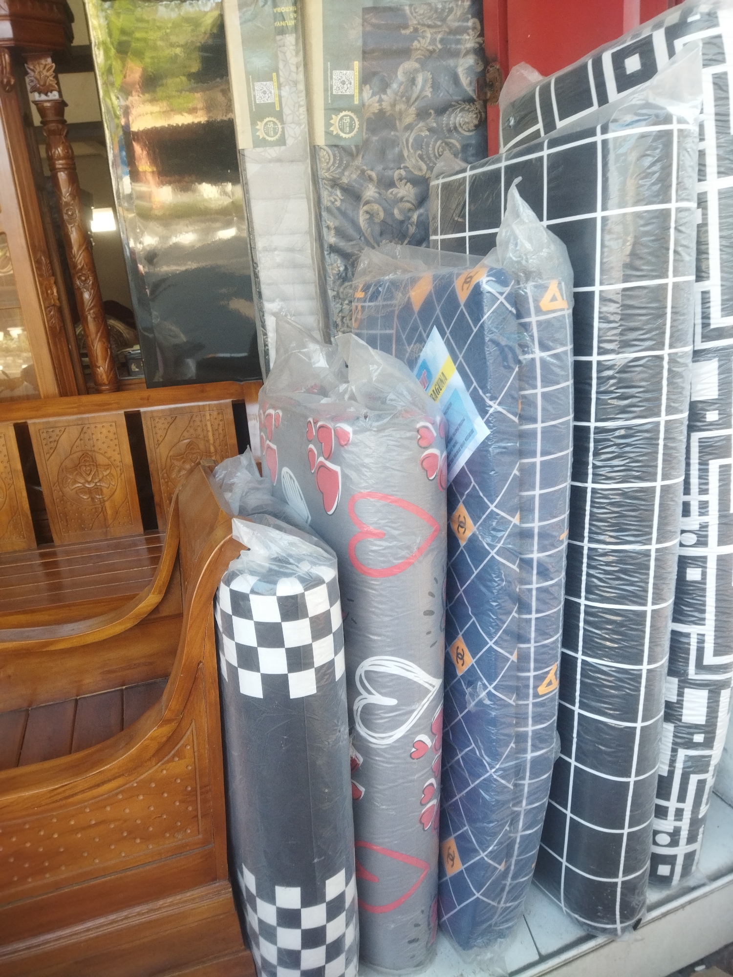 The image depicts a street-level view of a shop or stall in Sukoharjo, Indonesia, displaying a variety of rolled-up mattresses or similar bedding products. The items are wrapped in clear plastic, revealing different patterns: one with a black and white checkerboard design, another with a gray background and red heart shapes, a third with a dark blue background and a diamond pattern featuring orange logos, and several more with black backgrounds and white grid patterns. To the left, a portion of a wooden furniture item, possibly a bench or bed frame with intricate carvings, is visible. The background shows more rolled items and a hint of a building interior or doorway. The scene suggests a retail environment where these products are being sold.
