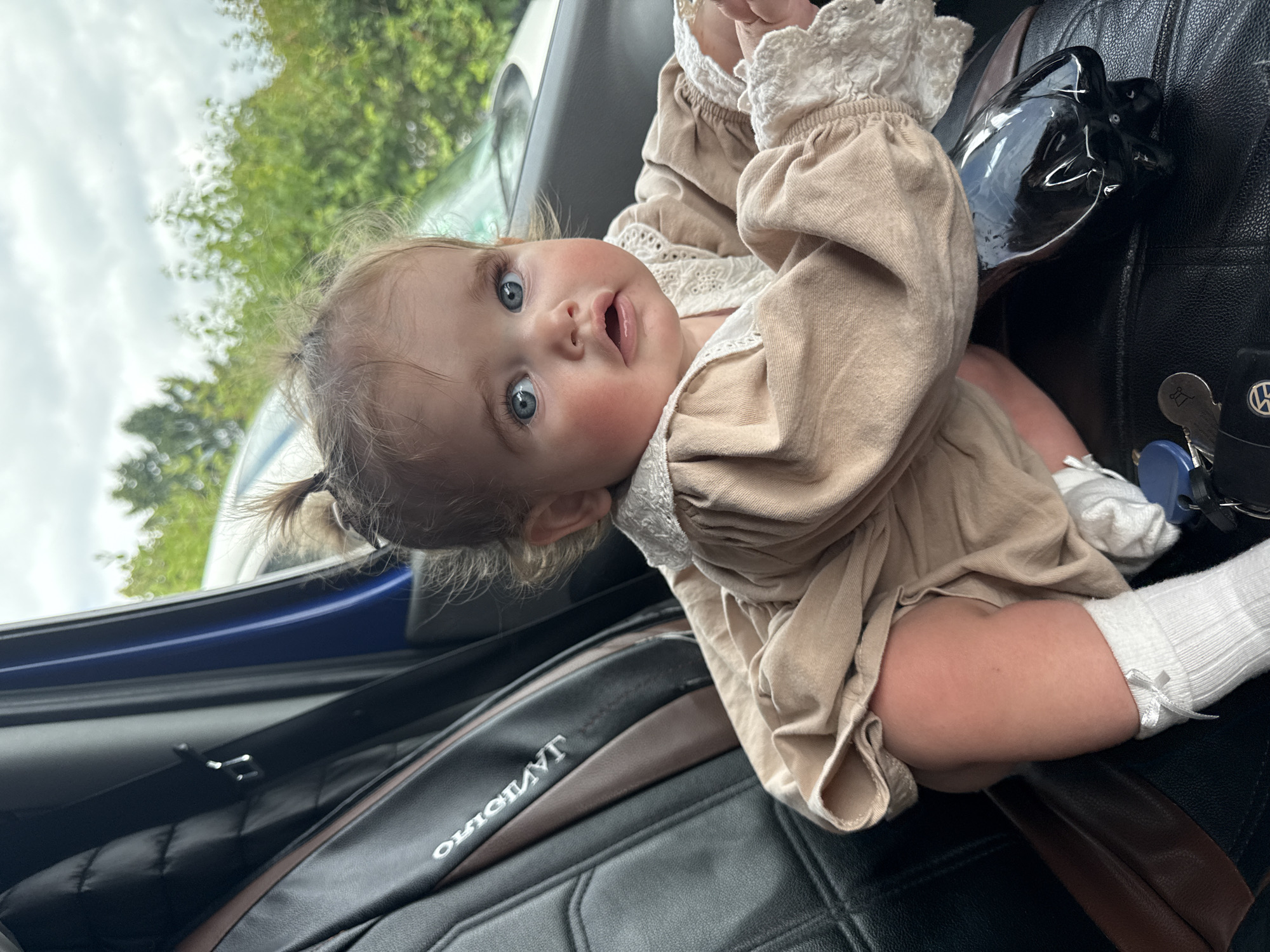 A baby girl with blue eyes and light brown hair is seated in a car. She is wearing a beige dress with lace trim and white socks with bows. The car has black leather seats with a brown accent. Outside the car, green trees and a cloudy sky are visible. The scene appears to be located in the city of Ulmen, Germany.