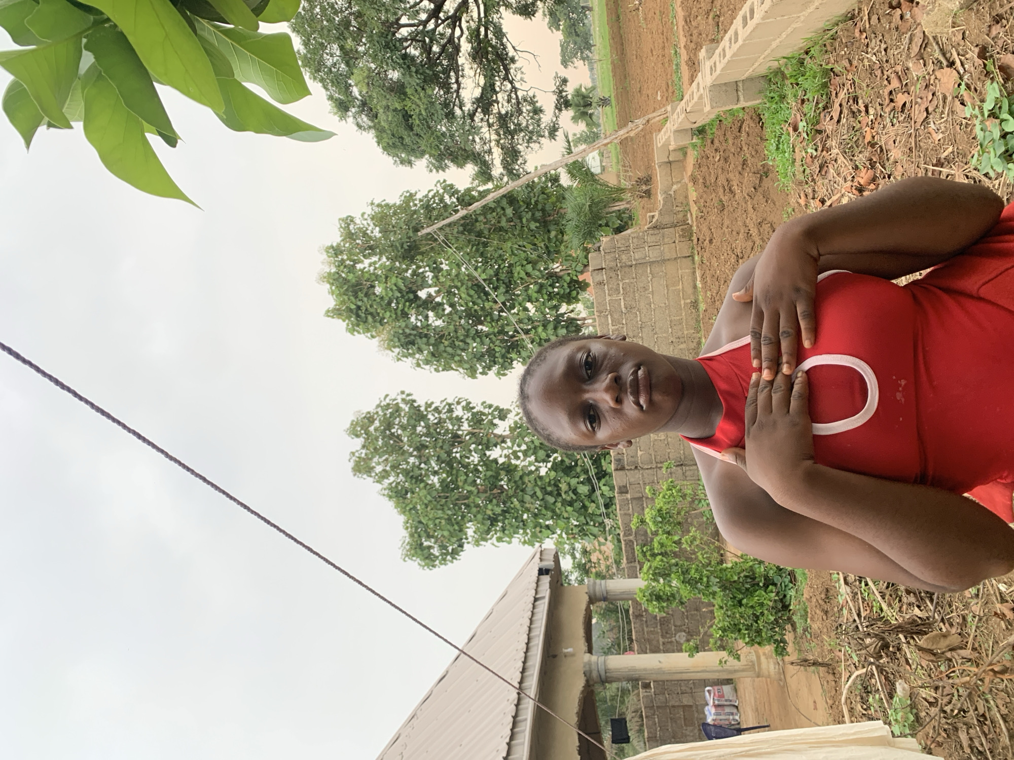 A medium shot angled upwards depicts a person of African descent in a red tank top with white trim, standing in an outdoor environment. The individual is looking toward the camera, with their hands crossed over their chest. The background consists of a landscape, including trees, bare soil, and a few structures. A powerline is visible against the overcast sky. The ground appears to be farmland or a yard with sparse vegetation and dry leaves. The location is indicated as Agban, Nigeria.