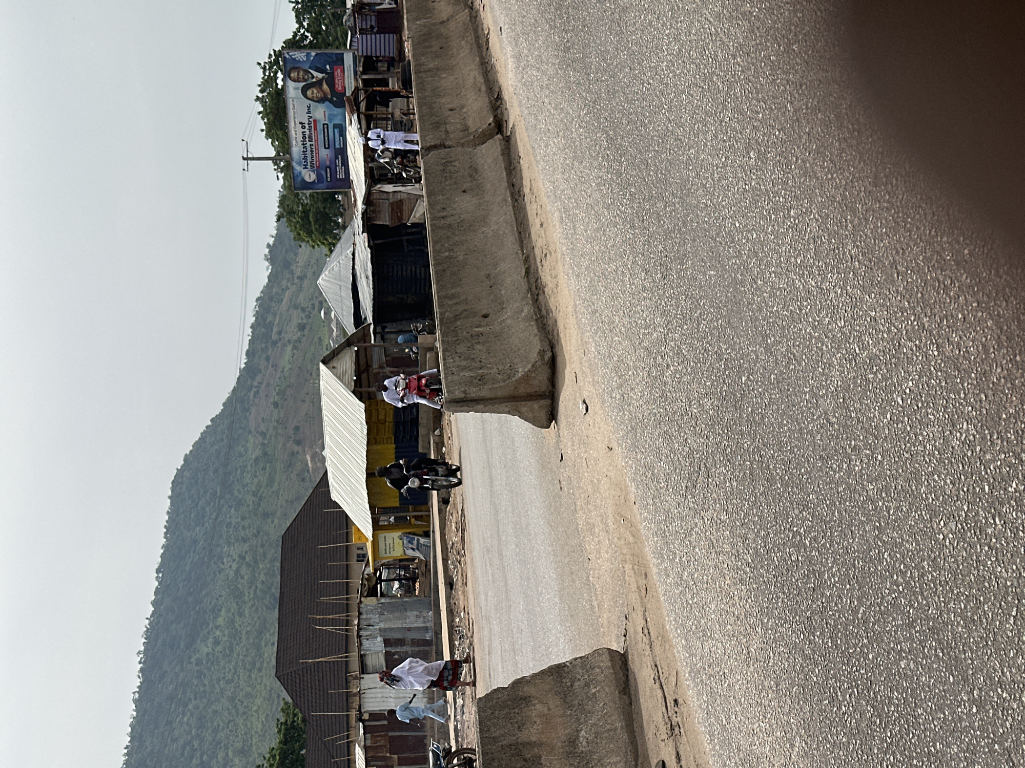 The image depicts a street scene in Lokoja, Nigeria. A paved road dominates the foreground, with a concrete barrier separating it from a row of informal shops and structures on the left. These shops appear to be constructed from corrugated metal and wood. Several individuals are visible, some wearing traditional white garments, and at least two are riding motorcycles. A billboard advertising a religious organization is visible in the distance. A large, green hill rises in the background, suggesting a terrain feature typical of the Lokoja area. The sky is bright and hazy.