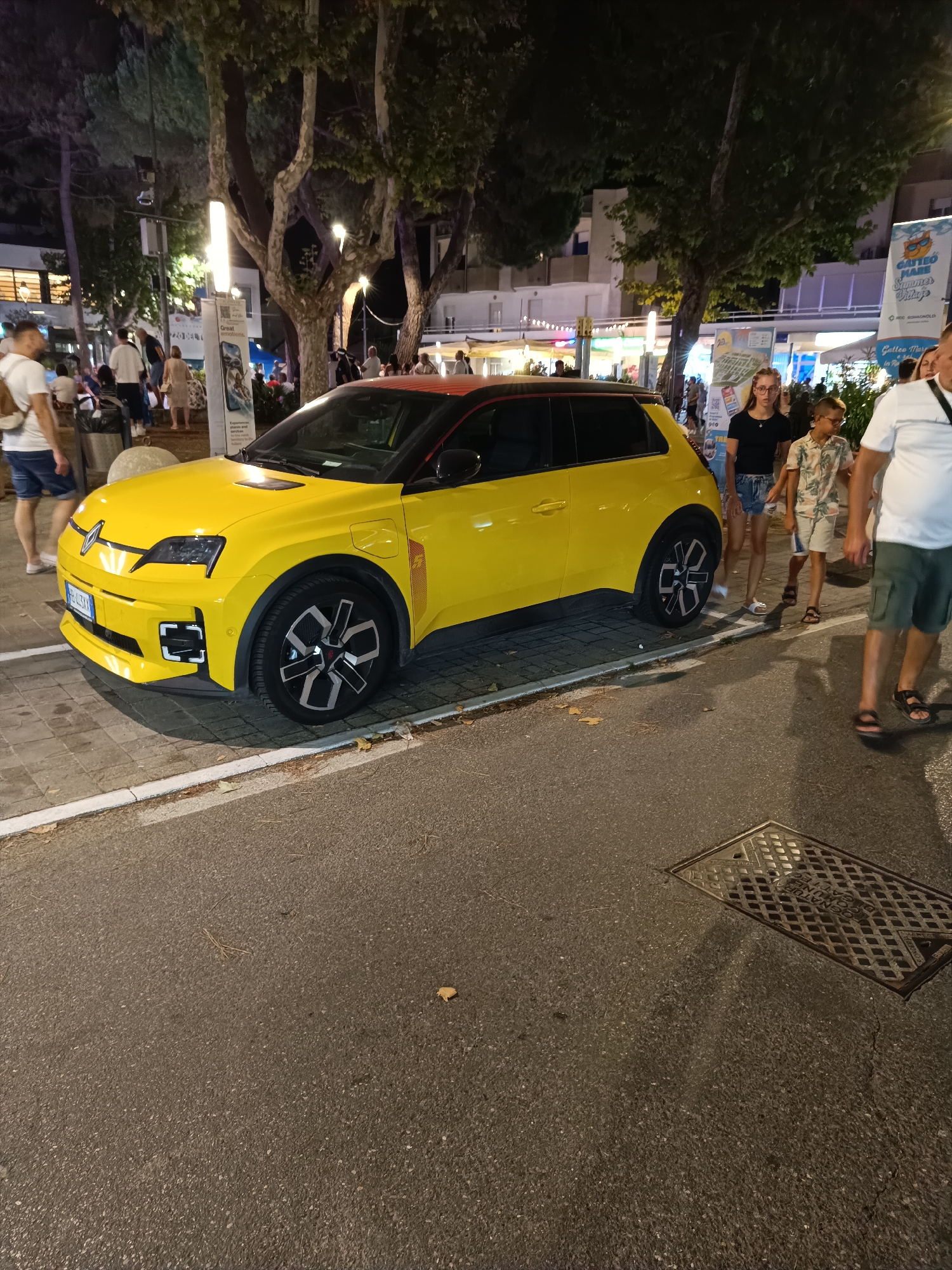 A yellow Renault 5 electric car is parked on a street in Gatteo, Italy, at night. The car has a black roof and red accents. In the background, people are walking on the sidewalk, and there are trees and buildings illuminated by streetlights. A man in a white t-shirt and shorts is walking towards the car from the left, while a woman and a boy in a floral shirt are walking away from the car on the right. The street is paved, and there is a sewer grate in the foreground. The scene appears to be a lively street with shops and restaurants.