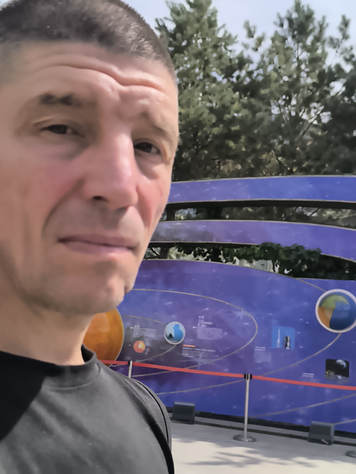 A man with a buzzed haircut and a black shirt is taking a selfie with a large, blue, planetarium-themed display in the background. The display features illustrations of planets and space, with a red rope barrier in front of it. The scene appears to be outdoors, with trees visible behind the display. The man's facial expression is neutral. The display shows a representation of the solar system with text and images of planets, rockets, and possibly satellites. The overall lighting suggests it is daytime. The background elements suggest this may be a public exhibition or science center.