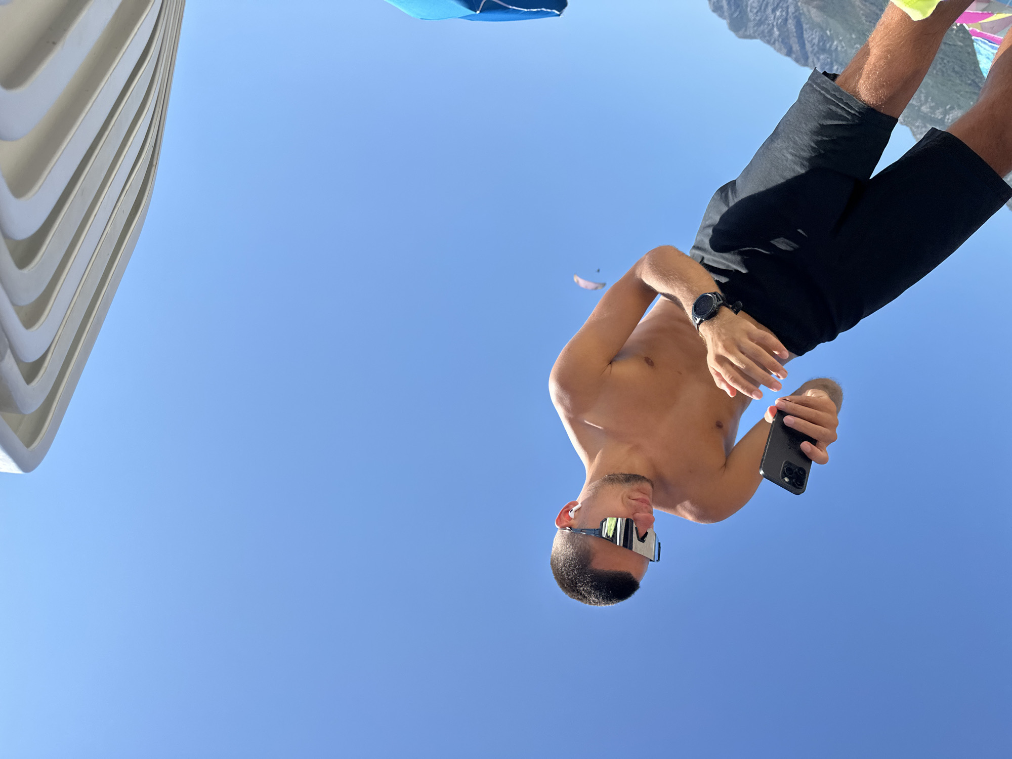 A young man, shirtless and wearing black shorts and sunglasses, is looking down at his phone. He is in an outdoor setting with a clear blue sky. In the background, a mountain range is visible, and there is a paraglider in the distance. A portion of a white, ribbed structure is in the upper right corner of the frame, and a blue umbrella or fabric is partially visible on the far right. The man's right wrist has a black smartwatch. The image appears to have been taken from a low angle, looking upwards.
