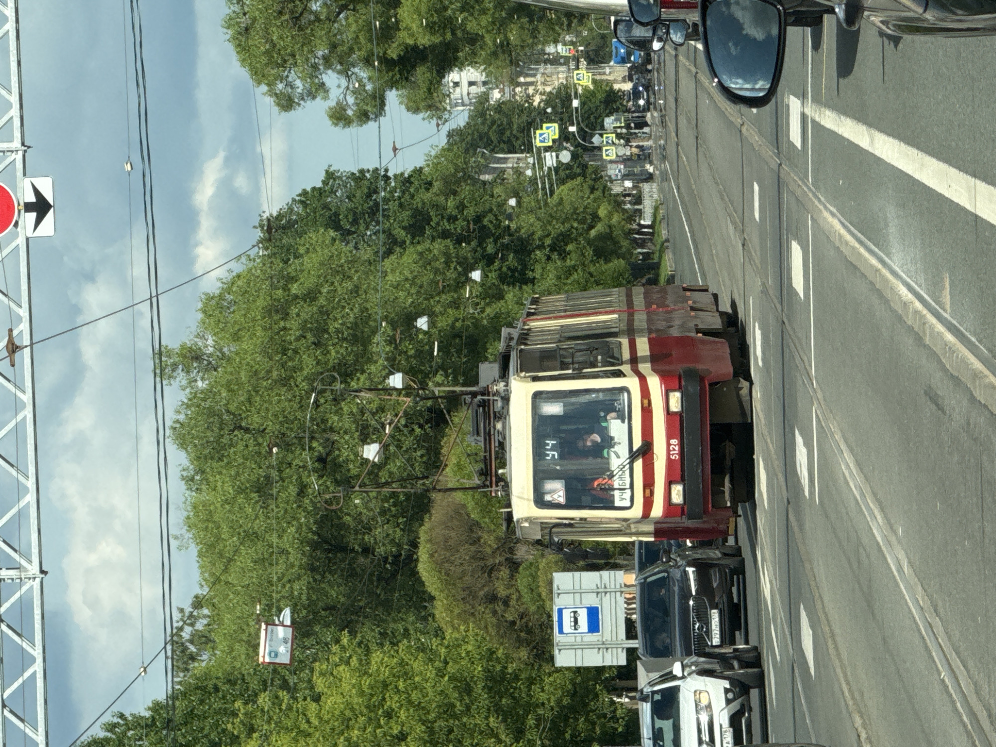 A street scene in Saint Petersburg, Russia, features a red and cream-colored tram numbered 5128, identified as route 44, proceeding along tram tracks on a paved road. The tram, with an operator visible inside, is surrounded by other vehicles, including a white SUV, a black Volvo SUV, and other cars. In the background, lush green trees line the street, and various traffic signals and signs are visible, including a bus stop sign. The sky above is cloudy, with patches of blue. The architectural style of the buildings in the distance suggests a historical urban setting.