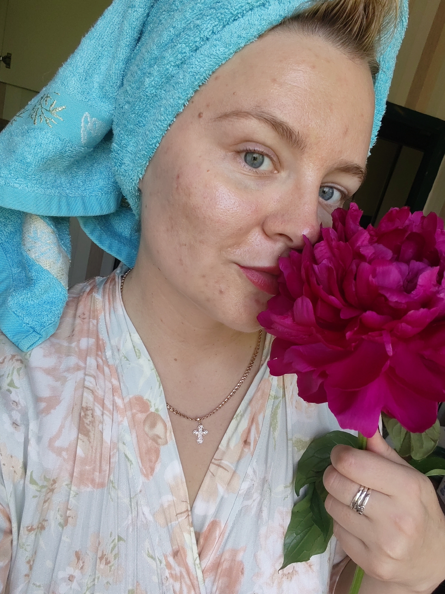 The image shows a close-up, selfie-style shot of a woman holding a large, dark pink peony flower. The woman is wearing a light blue towel wrapped around her head and a floral patterned robe. She has light blue eyes and her skin shows some redness and blemishes. She is also wearing a gold chain necklace with a cross pendant and two silver rings on her right hand. The background is blurred and appears to be indoors, possibly a domestic setting. There is no discernible information in the image to suggest a specific location within the city of Izhevsk, Russia.
